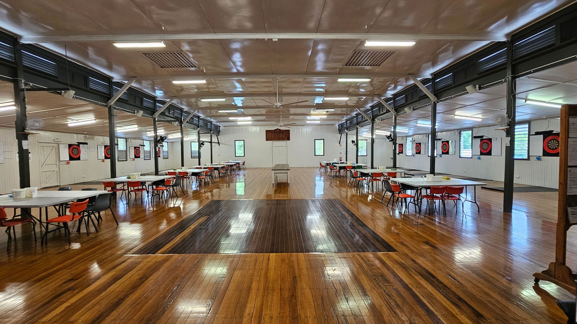 A spacious hall with a polished wooden floor and a white ceiling featuring fluorescent lights and ceiling fans. On the left, there are tables with red chairs arranged in rows. The right side has a notice board with various papers pinned to it. The back wall has dartboards and windows with green views outside. A long table is centred in the room. A spacious hall with a polished wooden floor and a white ceiling featuring fluorescent lights and ceiling fans. On the left, there are tables with red chairs arranged in rows. The right side has a notice board with various papers pinned to it. The back wall has dartboards and windows with green views outside. A long table is centred in the room.