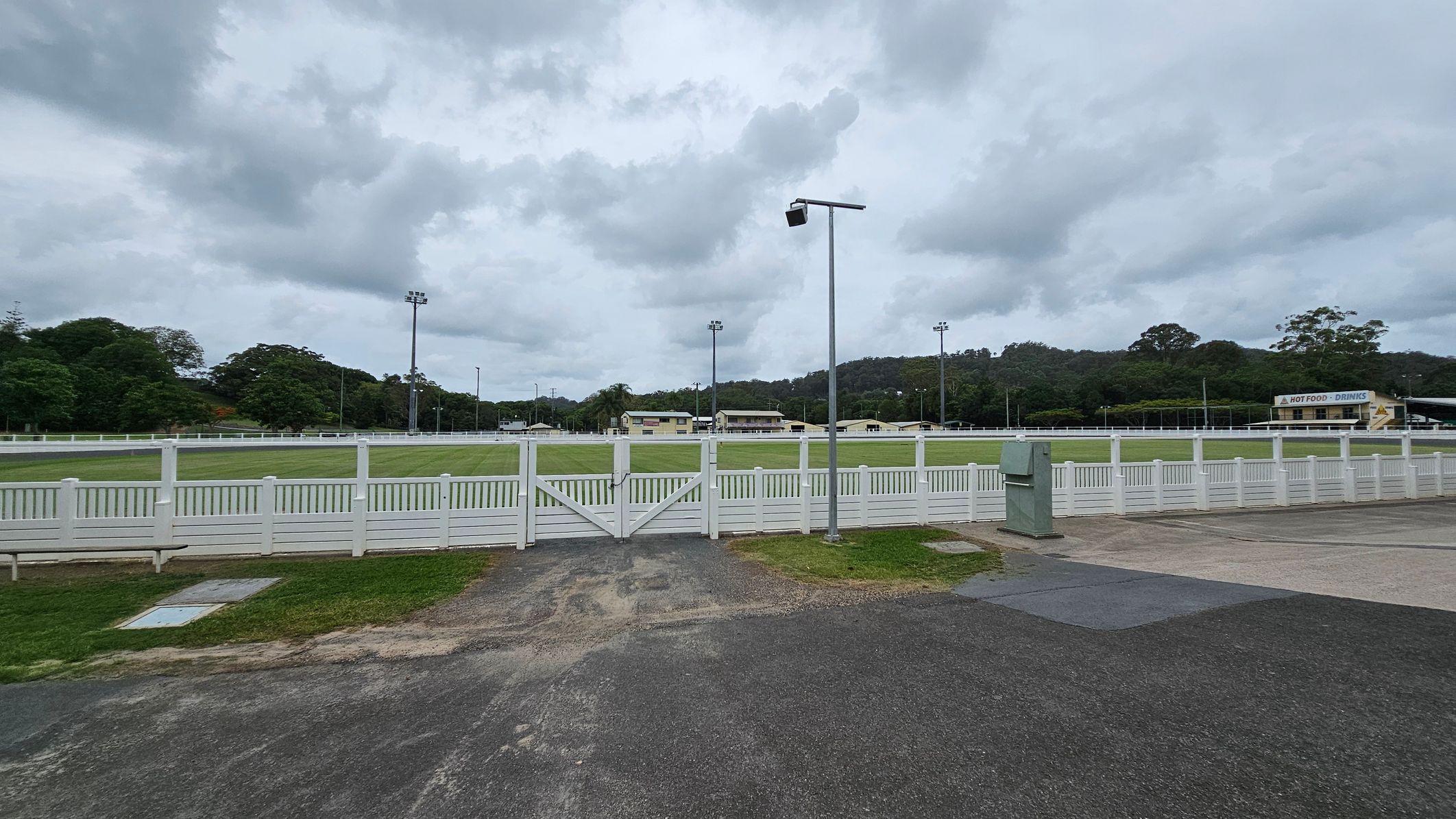 A sports field with a white fence runs across the middle, with a green lawn inside. Several floodlights are positioned around the field. In the background, there are buildings and trees on a hill. On the right, a yellow building with a sign for toilets is visible. The ground is paved with asphalt and concrete, and a bin is near the fence. The sky is overcast. A sports field with a white fence runs across the middle, with a green lawn inside. Several floodlights are positioned around the field. In the background, there are buildings and trees on a hill. On the right, a yellow building with a sign for toilets is visible. The ground is paved with asphalt and concrete, and a bin is near the fence. The sky is overcast.