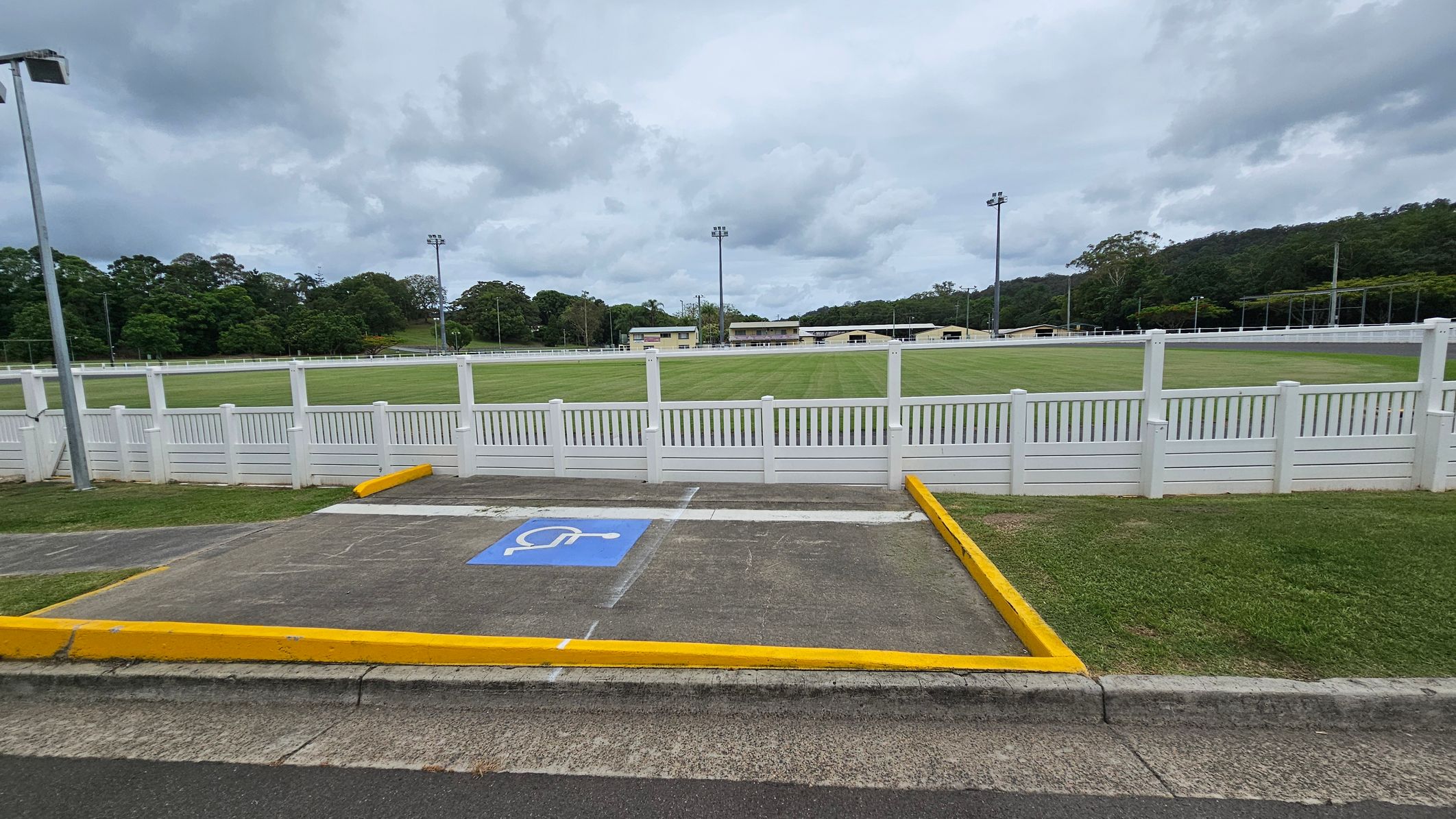A grassy sports field is surrounded by a white picket fence. In the foreground, there is a parking space marked with a blue disabled symbol, bordered by yellow lines. The background features trees and several tall floodlights. A few small buildings are visible beyond the field. The sky is overcast. A grassy sports field is surrounded by a white picket fence. In the foreground, there is a parking space marked with a blue disabled symbol, bordered by yellow lines. The background features trees and several tall floodlights. A few small buildings are visible beyond the field. The sky is overcast.