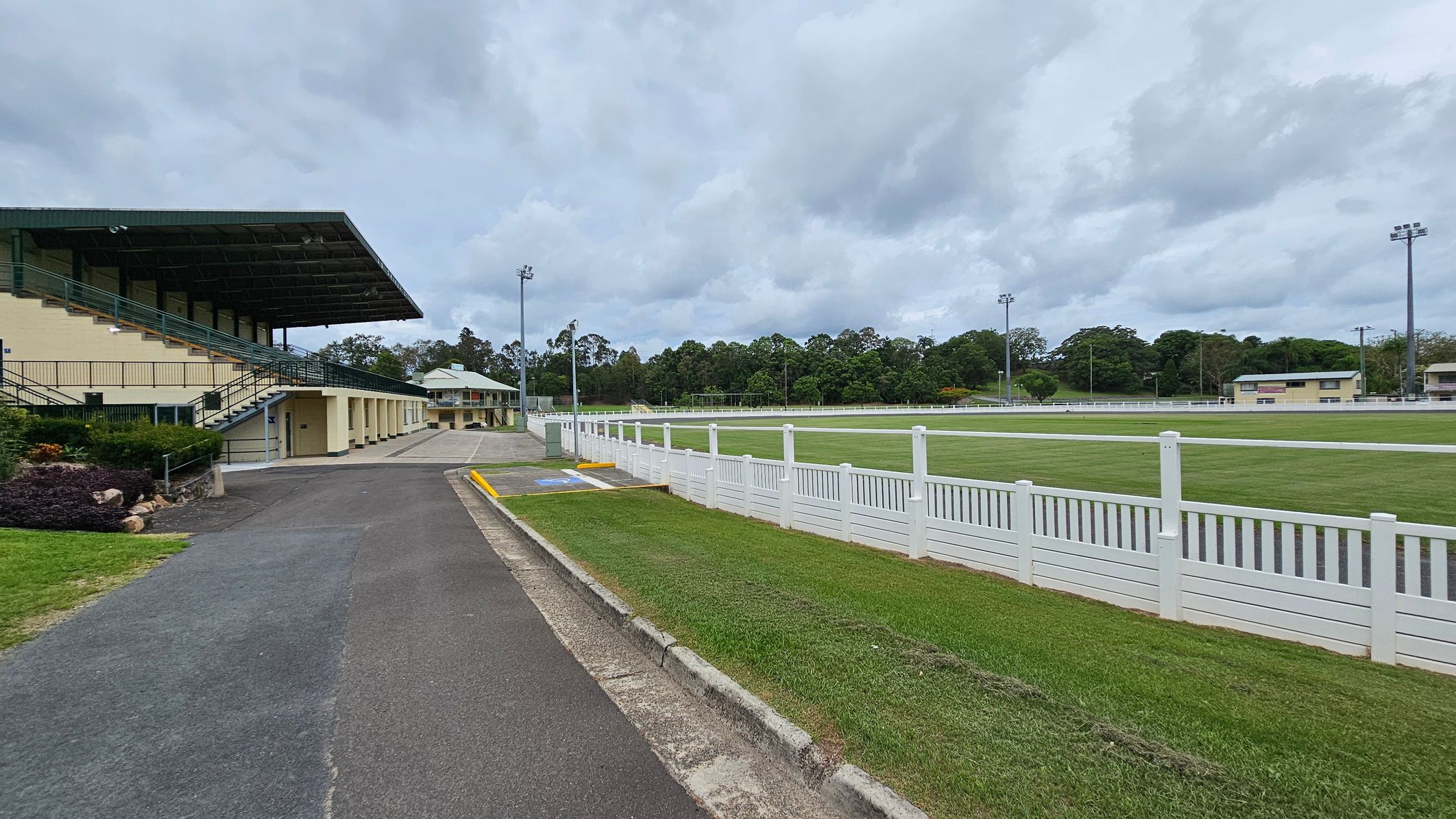 A sports field with a large grandstand on the left, featuring a green roof and multiple seating levels. A paved path runs alongside the grandstand, bordered by grass and a small garden. On the right, a white fence encloses a grassy field. In the background, there are trees and additional buildings, with tall floodlights visible around the field. The sky is overcast. A sports field with a large grandstand on the left, featuring a green roof and multiple seating levels. A paved path runs alongside the grandstand, bordered by grass and a small garden. On the right, a white fence encloses a grassy field. In the background, there are trees and additional buildings, with tall floodlights visible around the field. The sky is overcast.
