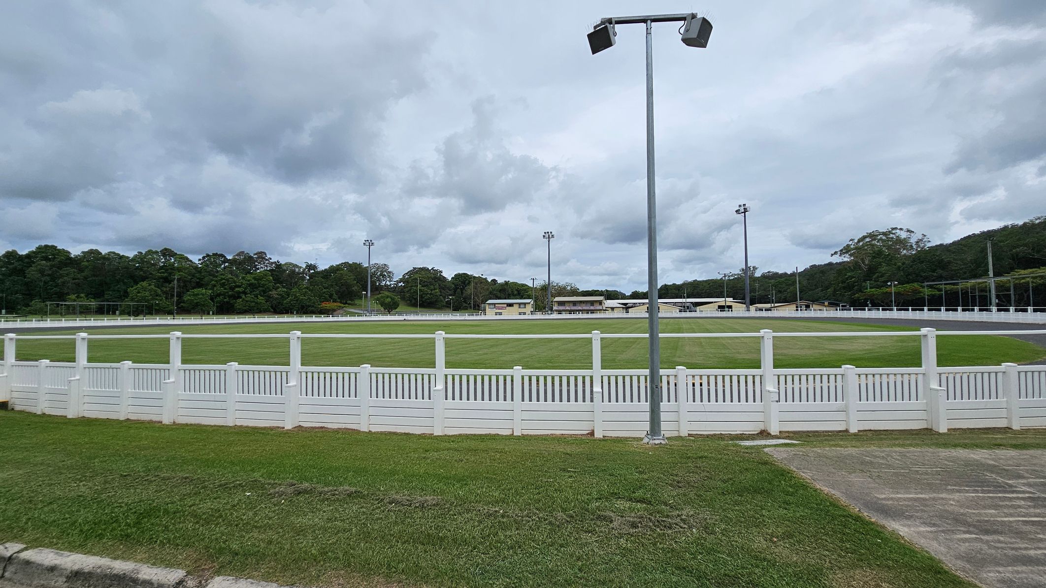 A grassy sports field is surrounded by a white picket fence. In the foreground, there is a patch of grass and a concrete path. A tall floodlight stands near the centre. In the background, there are several buildings and more floodlights. Dense trees line the horizon under a cloudy sky. A grassy sports field is surrounded by a white picket fence. In the foreground, there is a patch of grass and a concrete path. A tall floodlight stands near the centre. In the background, there are several buildings and more floodlights. Dense trees line the horizon under a cloudy sky.