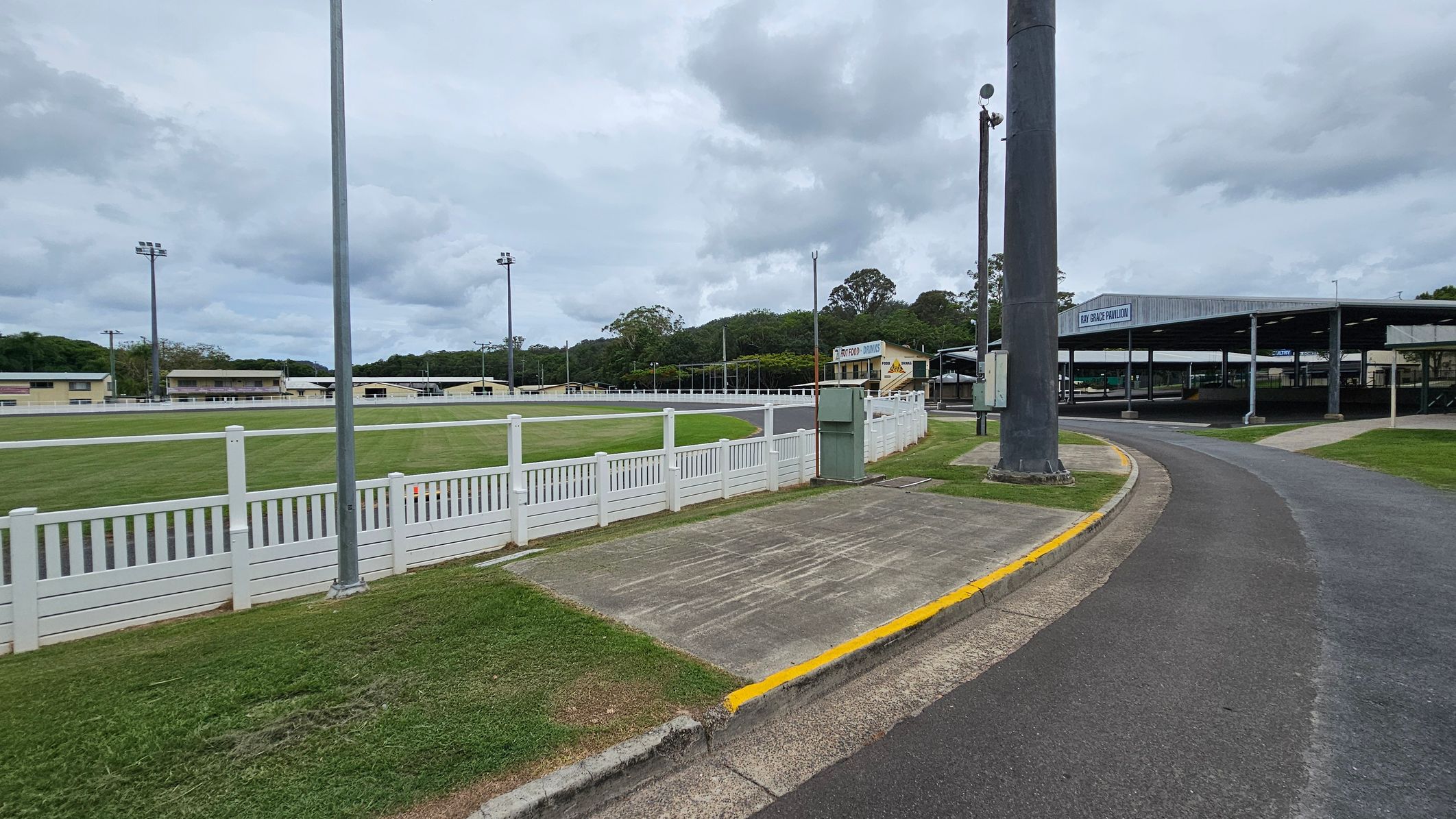 A paved road curves to the right, bordered by a grassy area and a white picket fence on the left. Beyond the fence is a green field with several tall floodlights. In the background, there are multiple low buildings, including one labelled "Bay Grace Pavilion." A large pole stands near the road, and trees are visible in the distance under a cloudy sky. A paved road curves to the right, bordered by a grassy area and a white picket fence on the left. Beyond the fence is a green field with several tall floodlights. In the background, there are multiple low buildings, including one labelled "Bay Grace Pavilion." A large pole stands near the road, and trees are visible in the distance under a cloudy sky.