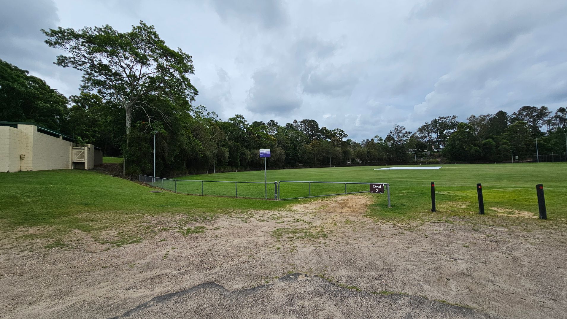 A grassy sports field with a sign reading "Oval 2" in the foreground. The field is surrounded by a low metal fence. On the left, there is a beige building with a small gate and a large tree nearby. The background features a line of dense trees. The ground in the foreground is a mix of grass and dirt, with several black bollards on the right side. A grassy sports field with a sign reading "Oval 2" in the foreground. The field is surrounded by a low metal fence. On the left, there is a beige building with a small gate and a large tree nearby. The background features a line of dense trees. The ground in the foreground is a mix of grass and dirt, with several black bollards on the right side.