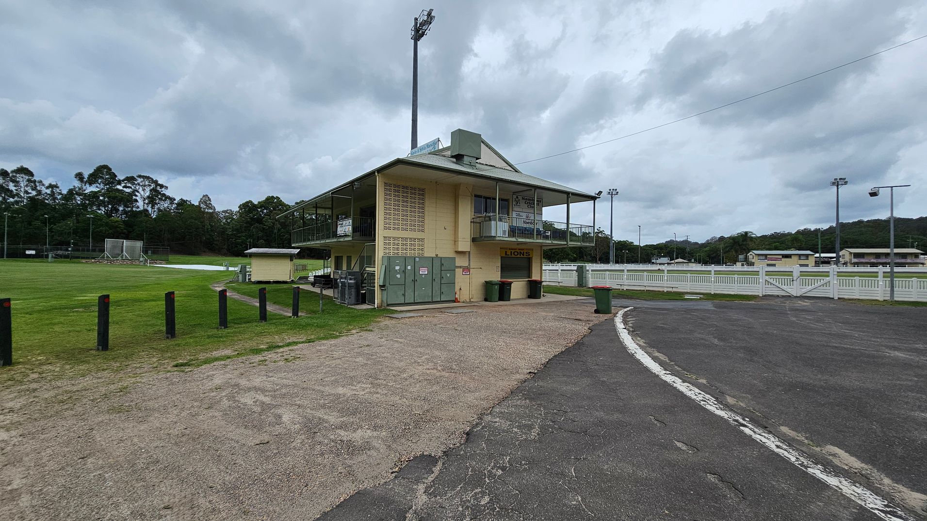 A sports field with a two-storey building on the right, featuring a sign that reads "LIONS" above a closed shutter. The building has a balcony with railings and several green bins nearby. The foreground has a mix of gravel and tarmac, leading to a curved road. On the left, there's a grassy field with black bollards and a netted practice area. In the background, there are trees and additional sports facilities, including floodlights and white fencing. A sports field with a two-storey building on the right, featuring a sign that reads "LIONS" above a closed shutter. The building has a balcony with railings and several green bins nearby. The foreground has a mix of gravel and tarmac, leading to a curved road. On the left, there's a grassy field with black bollards and a netted practice area. In the background, there are trees and additional sports facilities, including floodlights and white fencing.