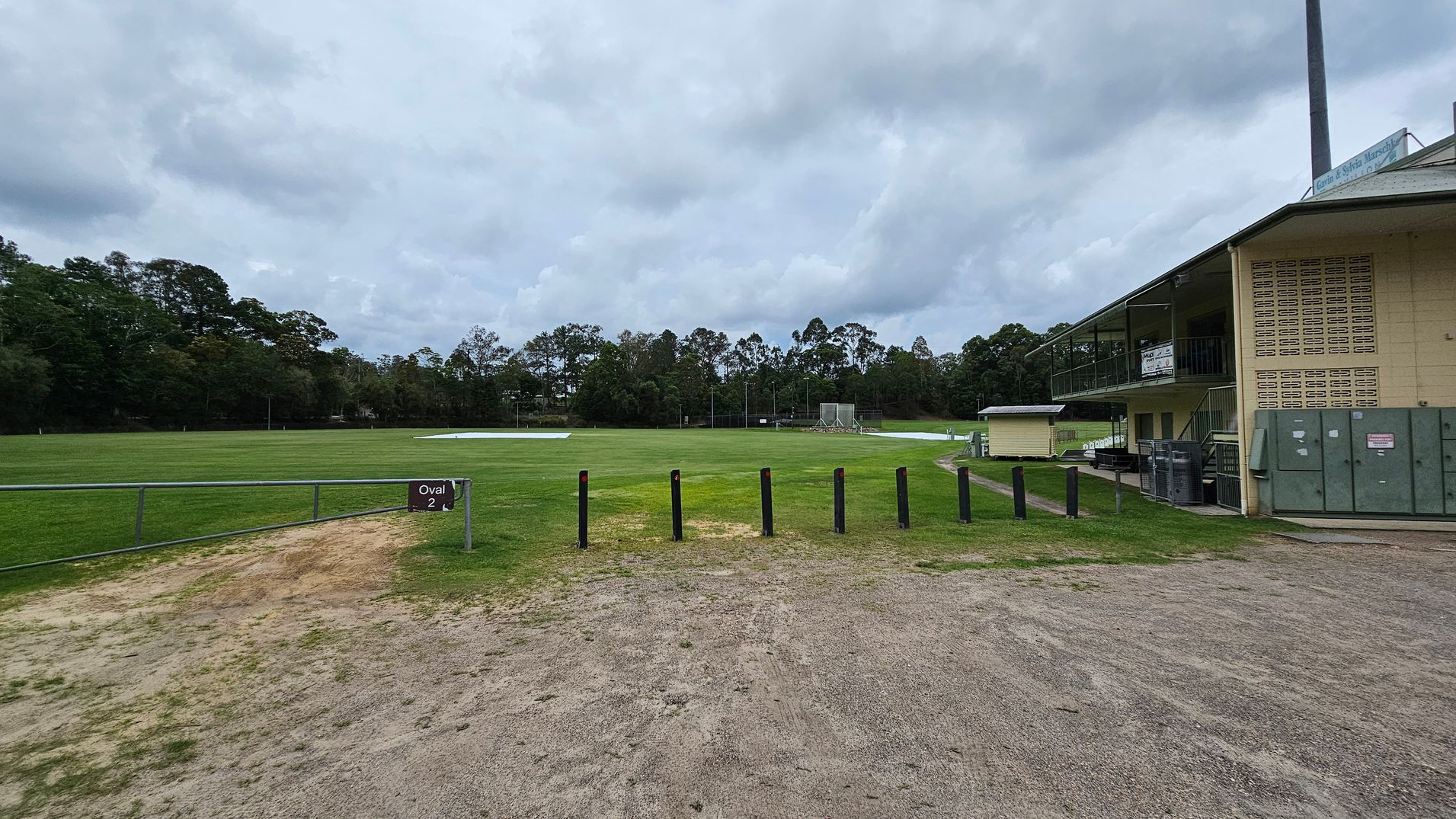 A grassy sports field labelled "Oval 2" is in the centre, surrounded by trees in the background. On the right, there is a two-storey building with a balcony and signs, alongside some utility boxes. The foreground has a mix of dirt and grass, with black bollards lining the edge of the field. A grassy sports field labelled "Oval 2" is in the centre, surrounded by trees in the background. On the right, there is a two-storey building with a balcony and signs, alongside some utility boxes. The foreground has a mix of dirt and grass, with black bollards lining the edge of the field.