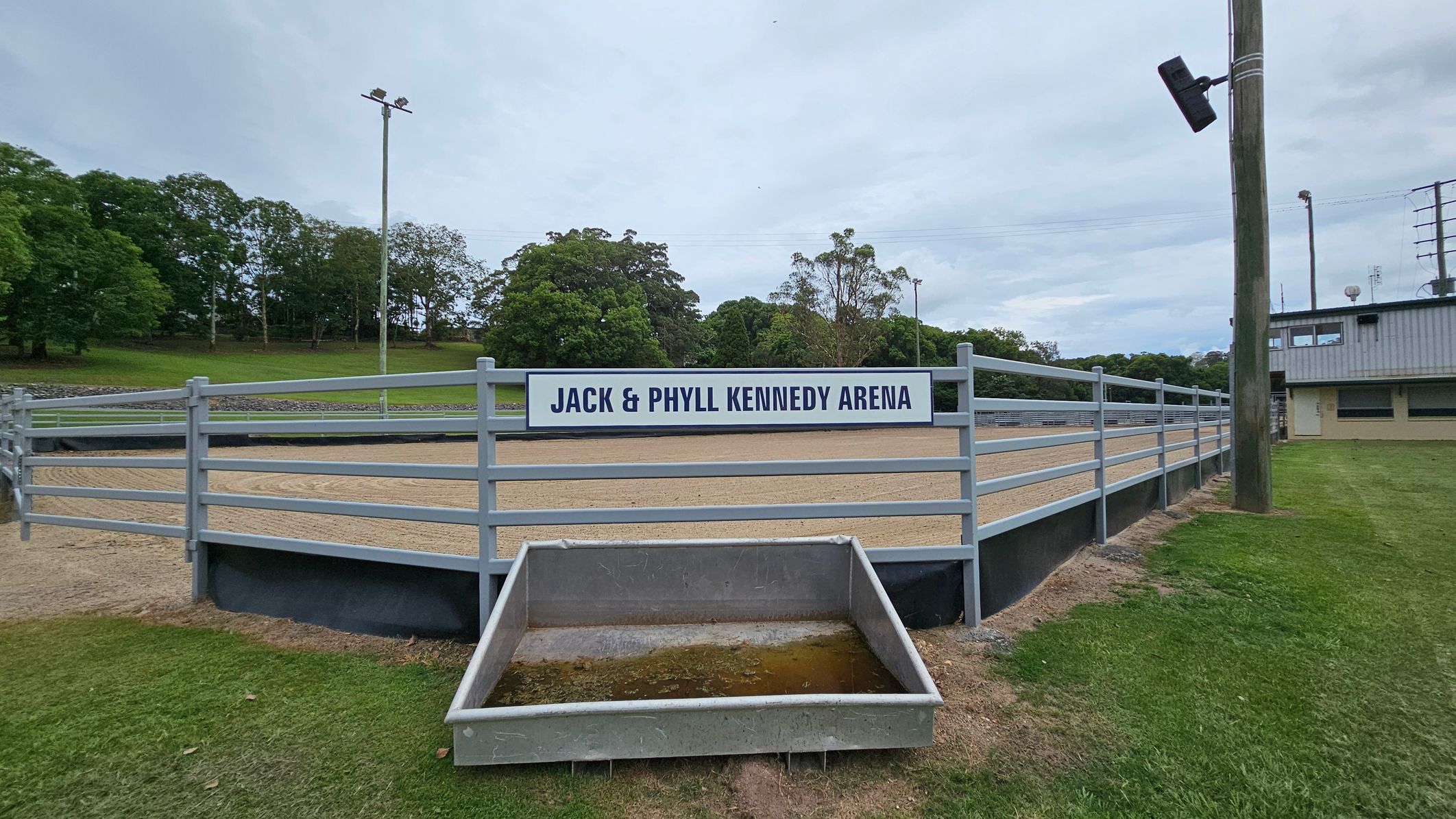 A fenced arena with a sign reading "Jack & Phyll Kennedy Arena" in the centre. The arena has a sandy surface and is surrounded by a metal railing. In the foreground, there is a rectangular concrete trough with some water. To the right, a wooden pole with a light fixture is visible, and a building with a corrugated metal roof is in the background. The left side features a grassy area with trees in the distance. A fenced arena with a sign reading "Jack & Phyll Kennedy Arena" in the centre. The arena has a sandy surface and is surrounded by a metal railing. In the foreground, there is a rectangular concrete trough with some water. To the right, a wooden pole with a light fixture is visible, and a building with a corrugated metal roof is in the background. The left side features a grassy area with trees in the distance.