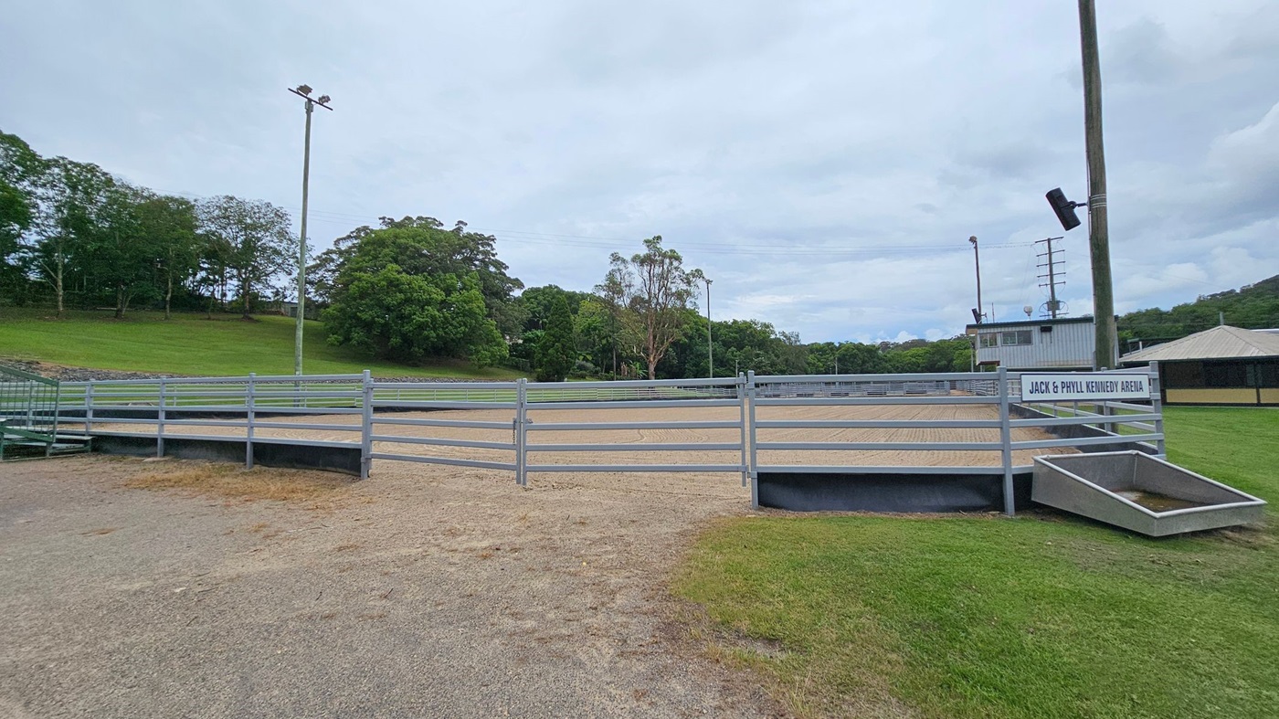 A large, sandy arena with visible rake patterns on the ground. On the left, there's a grassy embankment with dense green trees. On the right, a metal railing runs alongside a building with a corrugated metal roof and a sign partially visible. In the background, more trees and a few light poles are present. The sky is overcast. A large, sandy arena with visible rake patterns on the ground. On the left, there's a grassy embankment with dense green trees. On the right, a metal railing runs alongside a building with a corrugated metal roof and a sign partially visible. In the background, more trees and a few light poles are present. The sky is overcast.