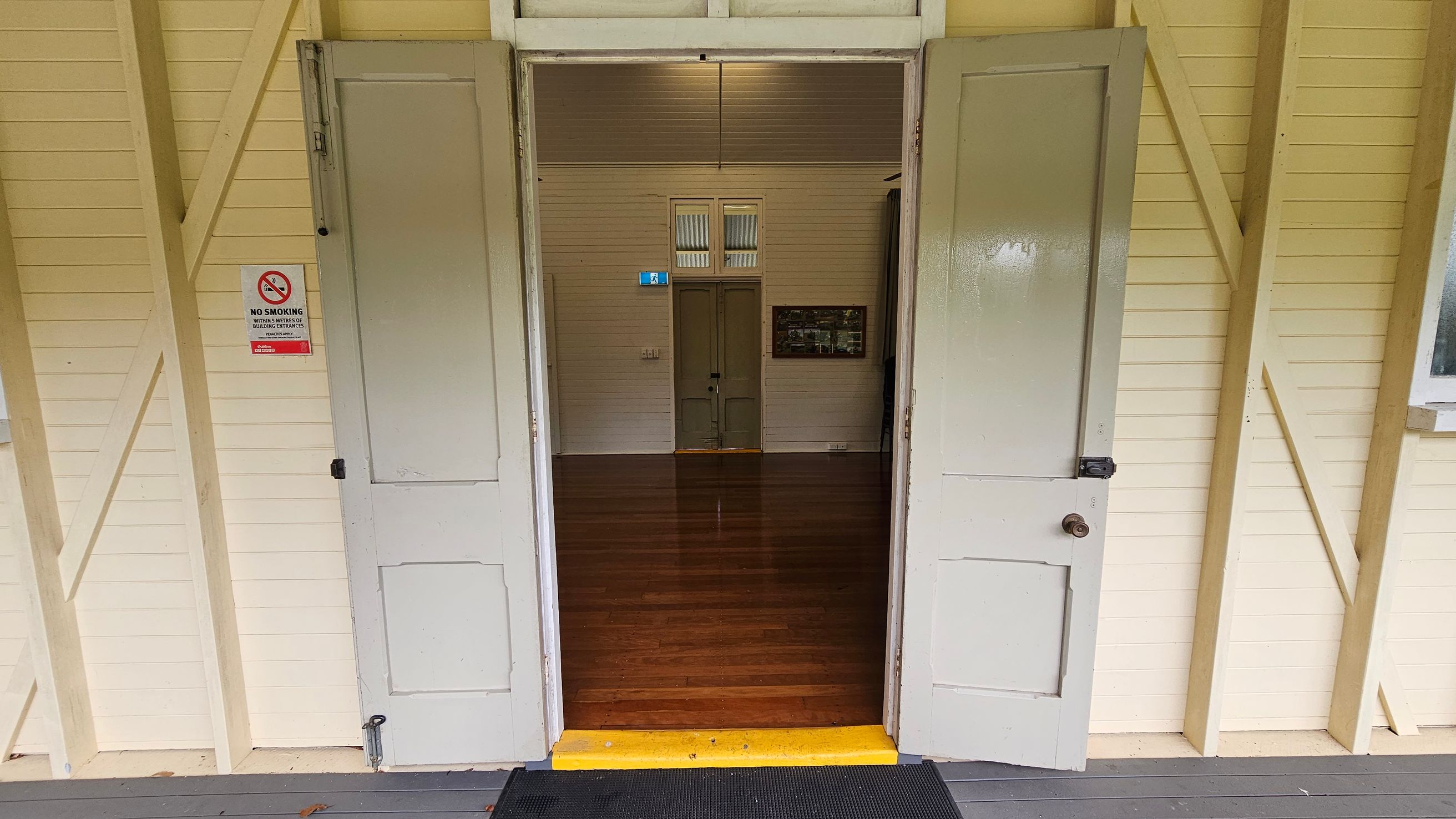 Open double doors lead into a room with polished wooden floors. The walls are cream-coloured with visible wooden beams. A "No Smoking" sign is on the left door. Inside, a closed door is visible at the far end, with a small window above it. A framed picture hangs on the right wall. The ceiling is wooden, matching the walls. A black mat and a yellow step are at the entrance. Open double doors lead into a room with polished wooden floors. The walls are cream-coloured with visible wooden beams. A "No Smoking" sign is on the left door. Inside, a closed door is visible at the far end, with a small window above it. A framed picture hangs on the right wall. The ceiling is wooden, matching the walls. A black mat and a yellow step are at the entrance.