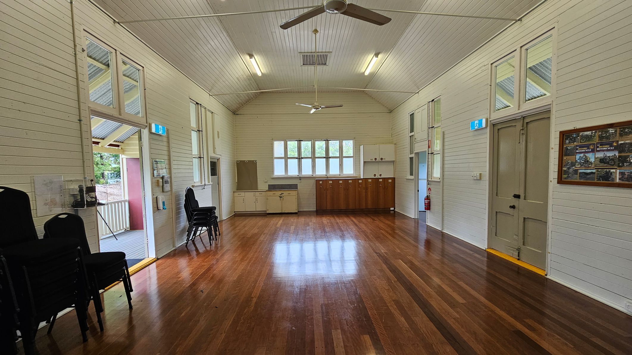 A spacious room with polished wooden floors and white panelled walls. On the left, there are stacked chairs and an open door leading to a porch. On the right, a closed door with a framed notice board nearby. The back wall features a row of windows above a series of cabinets, with a fire extinguisher to the side. The ceiling is high with two ceiling fans and fluorescent lights. A spacious room with polished wooden floors and white panelled walls. On the left, there are stacked chairs and an open door leading to a porch. On the right, a closed door with a framed notice board nearby. The back wall features a row of windows above a series of cabinets, with a fire extinguisher to the side. The ceiling is high with two ceiling fans and fluorescent lights.