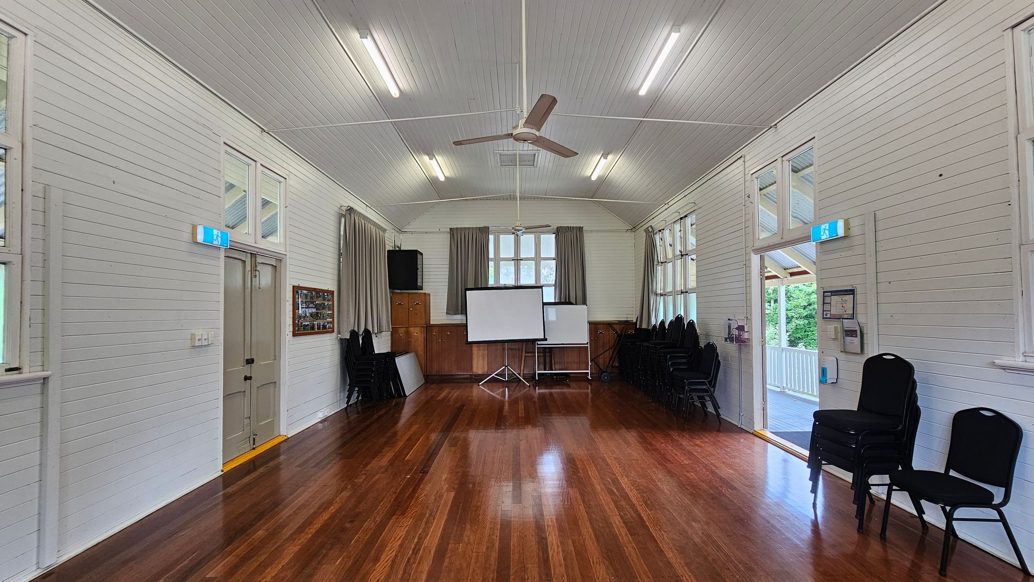 A hall with polished wooden floors and white panelled walls. In the centre, a projector screen stands in front of a window with curtains. On the left, a door with an exit sign above it, and a wall-mounted display. On the right, a stack of chairs and an open door leading to a balcony. The ceiling is vaulted with fluorescent lights and a ceiling fan. A hall with polished wooden floors and white panelled walls. In the centre, a projector screen stands in front of a window with curtains. On the left, a door with an exit sign above it, and a wall-mounted display. On the right, a stack of chairs and an open door leading to a balcony. The ceiling is vaulted with fluorescent lights and a ceiling fan.