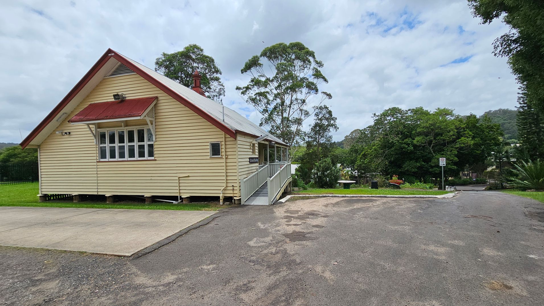 A small, single-storey wooden building with cream-coloured walls and a red roof is on the left. It has a window with an awning and a ramp leading to the entrance. The foreground features a paved area and a gravel path. In the background, there are tall trees and a grassy area with a bench and a signpost. The scene is surrounded by greenery, with hills visible in the distance. A small, single-storey wooden building with cream-coloured walls and a red roof is on the left. It has a window with an awning and a ramp leading to the entrance. The foreground features a paved area and a gravel path. In the background, there are tall trees and a grassy area with a bench and a signpost. The scene is surrounded by greenery, with hills visible in the distance.
