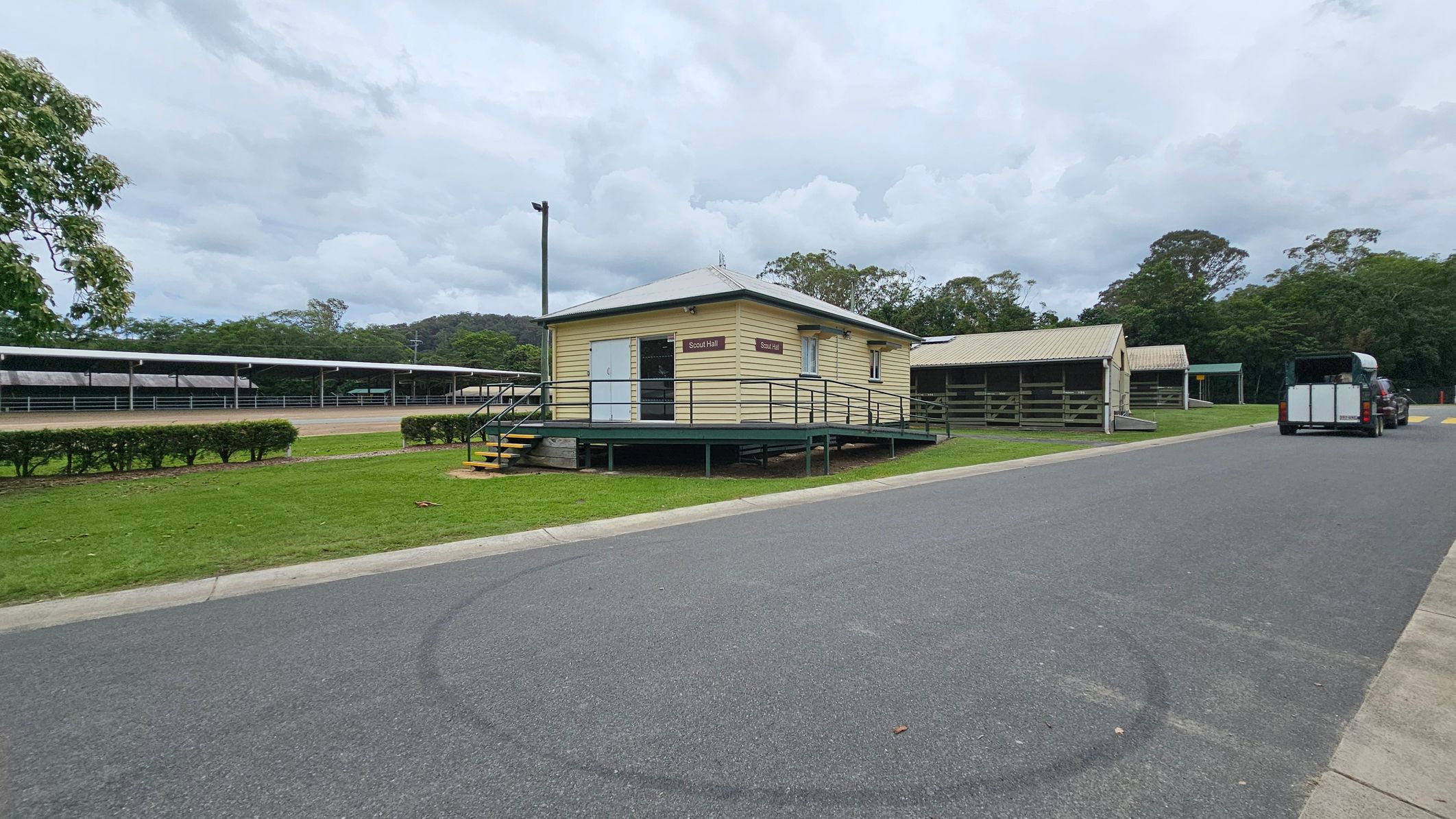 A small, elevated yellow building labelled "Scout Hall" is in the centre, with a ramp and steps leading up to it. To the right, a paved road runs alongside, with a vehicle towing a trailer parked further down. On the left, there are long, covered structures and a row of trimmed bushes. The background features dense green trees and a cloudy sky. The ground is a mix of grass and asphalt. A small, elevated yellow building labelled "Scout Hall" is in the centre, with a ramp and steps leading up to it. To the right, a paved road runs alongside, with a vehicle towing a trailer parked further down. On the left, there are long, covered structures and a row of trimmed bushes. The background features dense green trees and a cloudy sky. The ground is a mix of grass and asphalt.