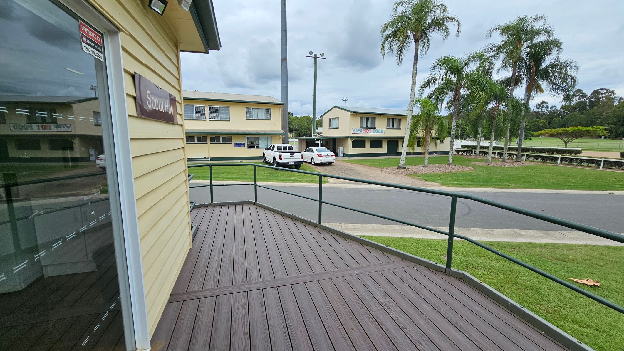 A view from a wooden deck with a green railing. On the left, a building with a sign reading "Scout Hall" is visible. In the background, there are two parked vehicles, a white pickup truck and a white car, in front of a yellow building with signs. To the right, a row of tall palm trees lines a grassy area. The scene includes a paved road and more greenery in the distance. A view from a wooden deck with a green railing. On the left, a building with a sign reading "Scout Hall" is visible. In the background, there are two parked vehicles, a white pickup truck and a white car, in front of a yellow building with signs. To the right, a row of tall palm trees lines a grassy area. The scene includes a paved road and more greenery in the distance.