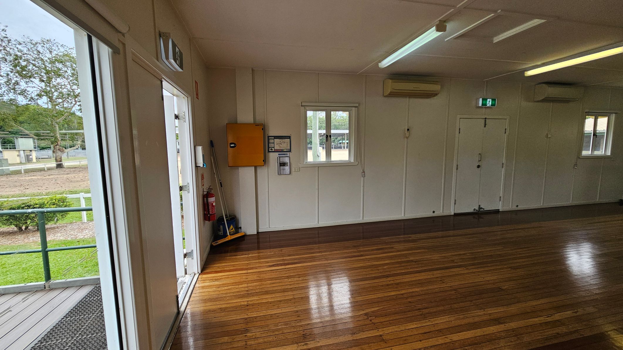 A room with polished wooden floors and white walls. On the left, there's an open door leading outside, showing a grassy area and a tree. Inside, a fire extinguisher and cleaning supplies are near the door. The back wall has a small window, a yellow cabinet, and an air conditioning unit. The ceiling has fluorescent lights, and there's an exit sign above a door on the right. A room with polished wooden floors and white walls. On the left, there's an open door leading outside, showing a grassy area and a tree. Inside, a fire extinguisher and cleaning supplies are near the door. The back wall has a small window, a yellow cabinet, and an air conditioning unit. The ceiling has fluorescent lights, and there's an exit sign above a door on the right.