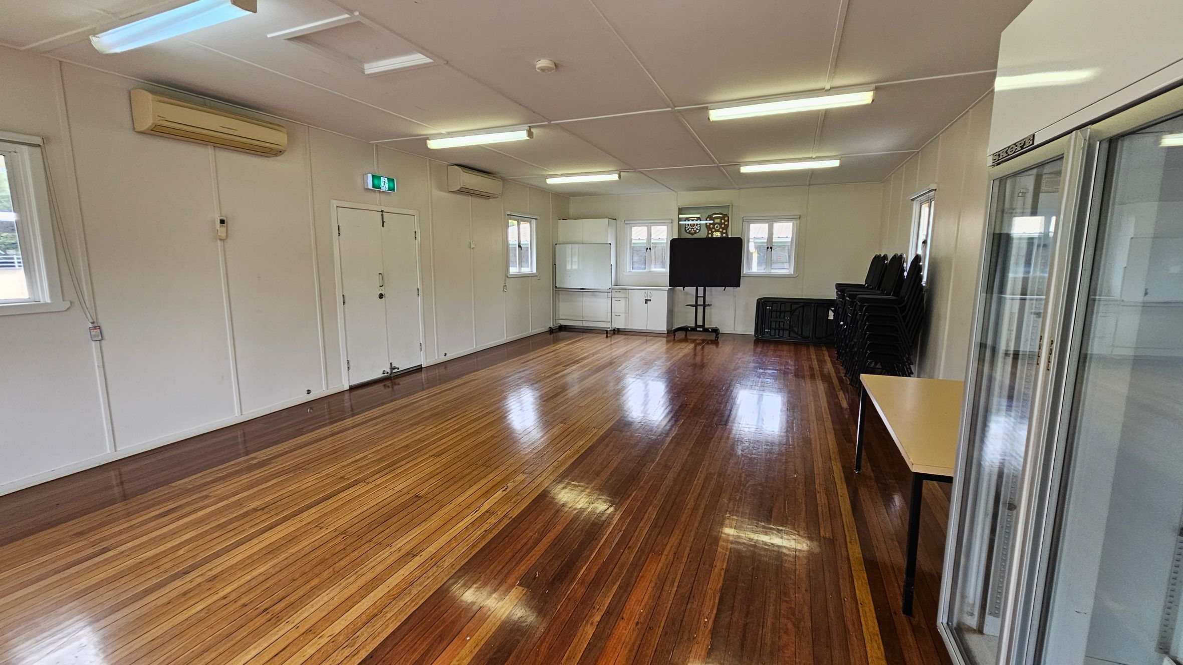 A spacious room with polished wooden floors and a white ceiling with fluorescent lights. On the left, there are white walls with air conditioning units and a double door. The back wall has windows, a white cabinet, and a black screen on a stand. On the right, there is a stack of black chairs, a folded table, and a glass-fronted refrigerator. A spacious room with polished wooden floors and a white ceiling with fluorescent lights. On the left, there are white walls with air conditioning units and a double door. The back wall has windows, a white cabinet, and a black screen on a stand. On the right, there is a stack of black chairs, a folded table, and a glass-fronted refrigerator.