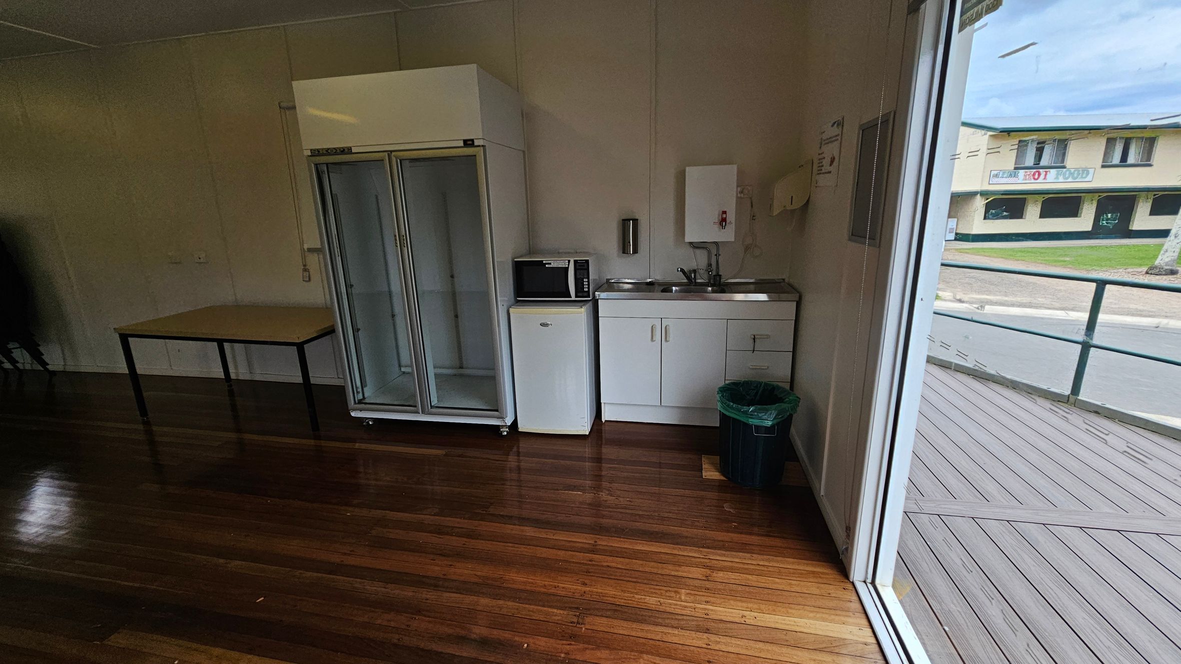 A small kitchenette with a wooden floor. On the left, there's a table and a large glass-fronted fridge. In the centre, a microwave sits atop a small fridge next to a sink with a cabinet below. A green bin is on the floor. On the right, a large window shows an outdoor deck and a view of a building across the street with a sign that reads "HOT FOOD". A small kitchenette with a wooden floor. On the left, there's a table and a large glass-fronted fridge. In the centre, a microwave sits atop a small fridge next to a sink with a cabinet below. A green bin is on the floor. On the right, a large window shows an outdoor deck and a view of a building across the street with a sign that reads "HOT FOOD".