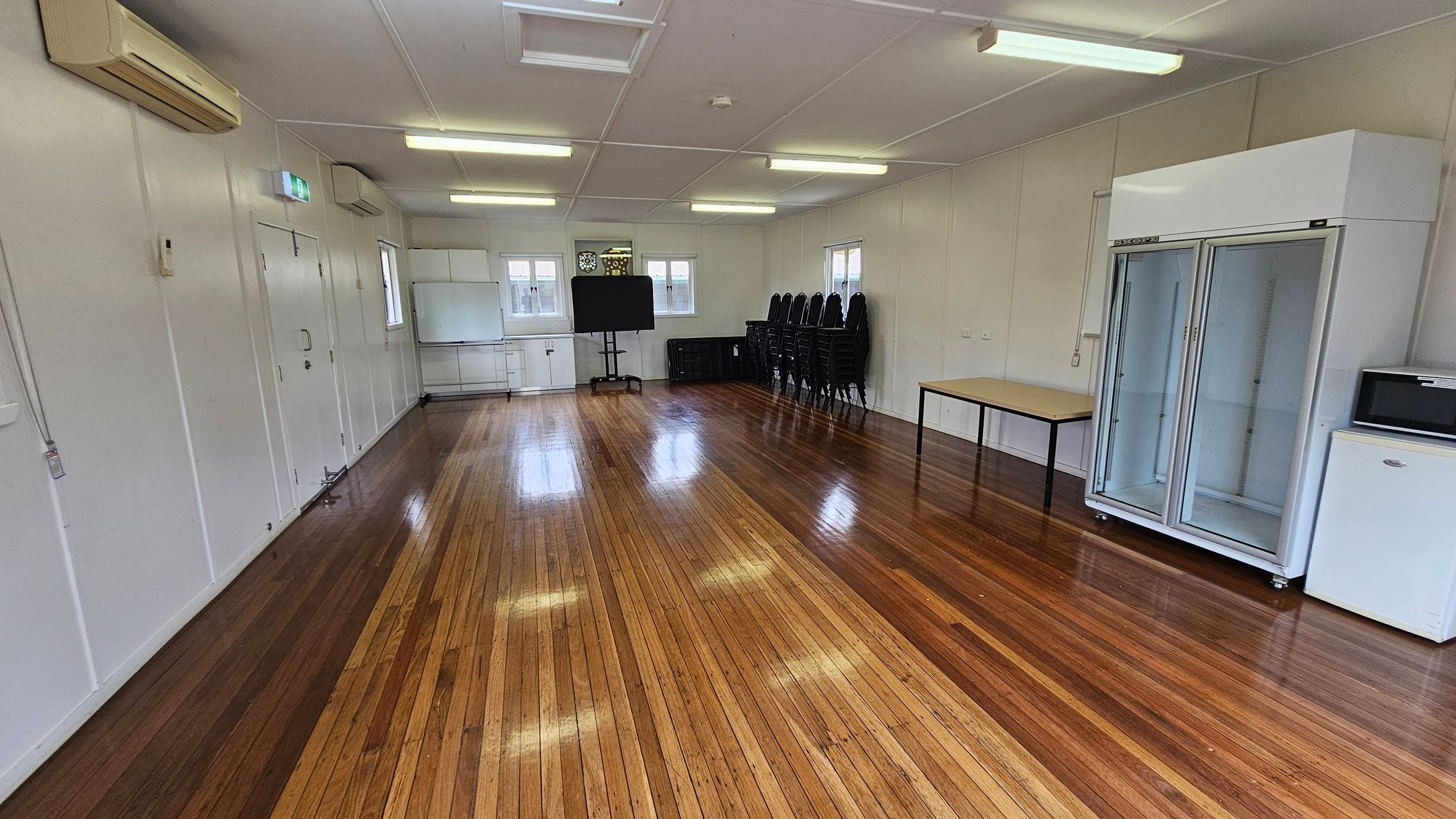 A spacious room with polished wooden floors and a white ceiling with fluorescent lights. On the left, there's an air conditioning unit and a door. In the middle, a blackboard and stacked chairs are against the wall. On the right, a large glass-fronted refrigerator, a table, and a small white fridge are visible. Windows are present on the far wall, allowing natural light in. A spacious room with polished wooden floors and a white ceiling with fluorescent lights. On the left, there's an air conditioning unit and a door. In the middle, a blackboard and stacked chairs are against the wall. On the right, a large glass-fronted refrigerator, a table, and a small white fridge are visible. Windows are present on the far wall, allowing natural light in.