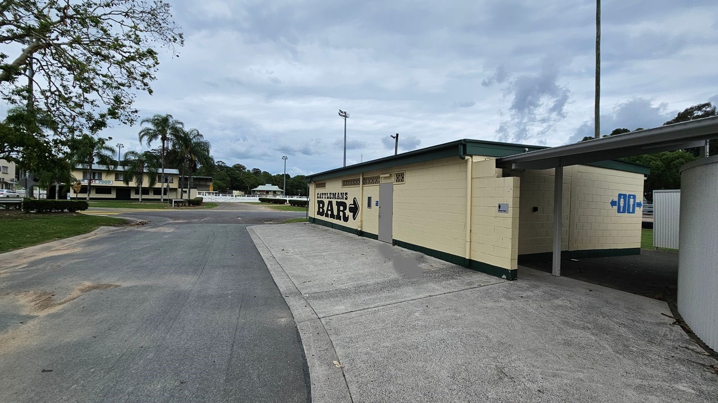 A single-storey, cream-coloured building with green trims is on the right, displaying the text "Cattleman’s Bar" and directional signage. Adjacent to it, a paved concrete area transitions to a tarmac driveway. On the left, a tree with sparse branches is over a grassy area, and in the background, a multi-storey building with palm trees. The foreground features a slightly uneven tarmac surface, while tall light poles rise in the middle ground. A single-storey, cream-coloured building with green trims is on the right, displaying the text "Cattleman’s Bar" and directional signage. Adjacent to it, a paved concrete area transitions to a tarmac driveway. On the left, a tree with sparse branches is over a grassy area, and in the background, a multi-storey building with palm trees. The foreground features a slightly uneven tarmac surface, while tall light poles rise in the middle ground.