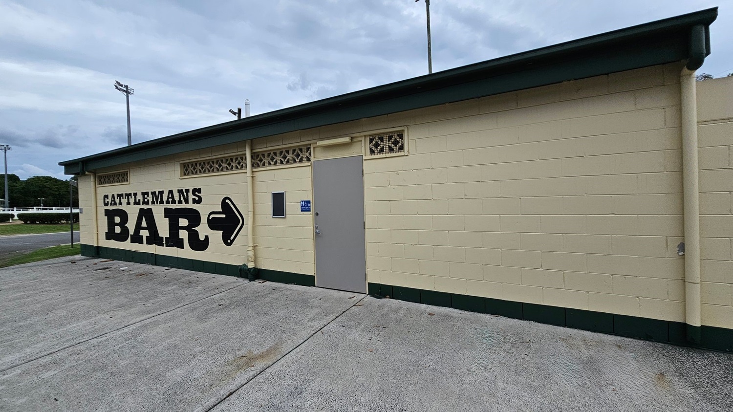 A cream-coloured single-storey building with green trims is on the right, featuring "Cattleman’s Bar" text and an arrow pointing right. A grey door and a ventilation block sit in the centre, with concrete paving on the foreground. The left shows part of a grassy area, a road, and some trimmed bushes. Tall light poles rise in the background. The ground is a concrete surface with scattered leaves, while the ceiling is dark green overhang roofing. A cream-coloured single-storey building with green trims is on the right, featuring "Cattleman’s Bar" text and an arrow pointing right. A grey door and a ventilation block sit in the centre, with concrete paving on the foreground. The left shows part of a grassy area, a road, and some trimmed bushes. Tall light poles rise in the background. The ground is a concrete surface with scattered leaves, while the ceiling is dark green overhang roofing.