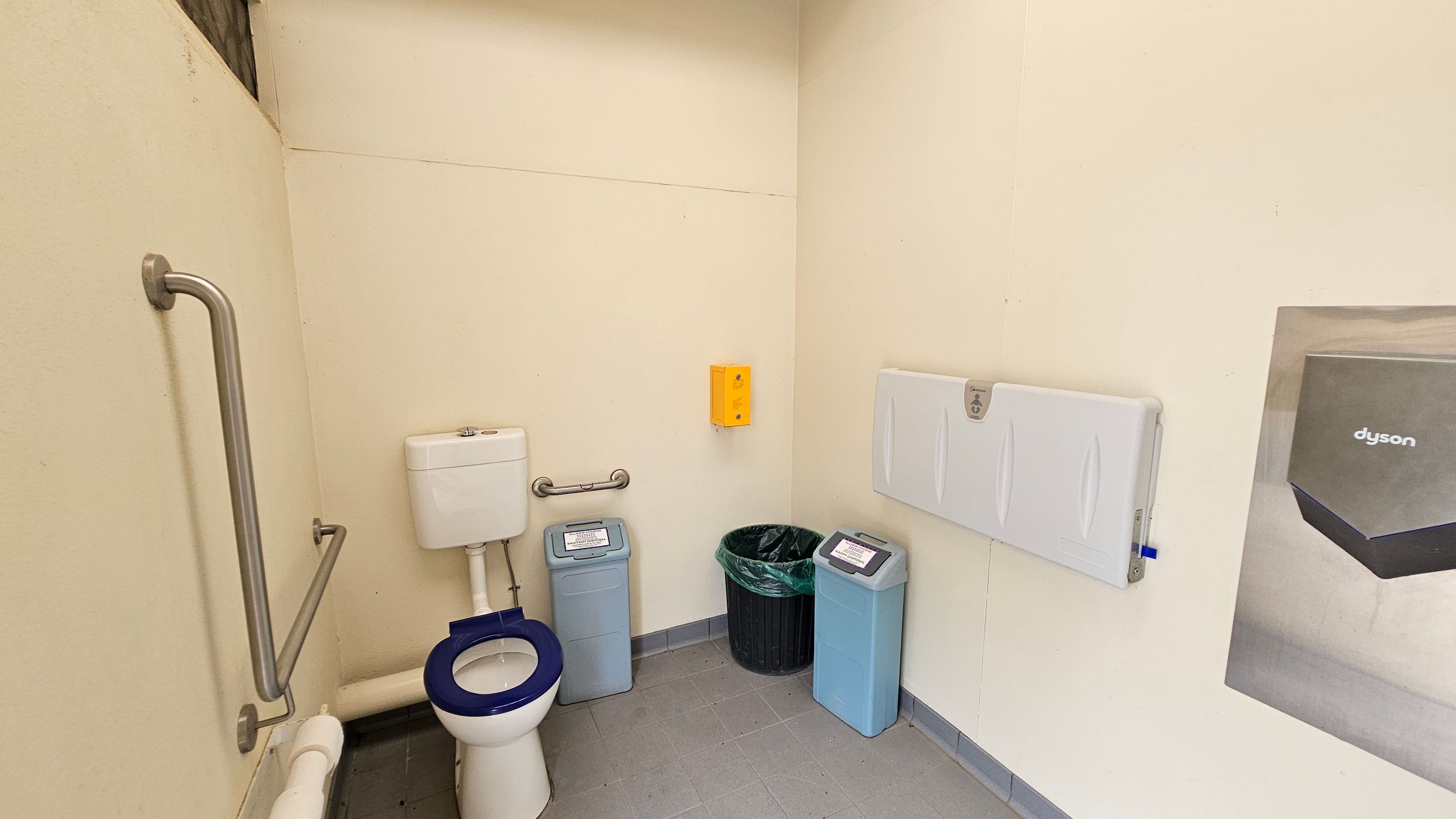 A white toilet with a blue seat is positioned on the left against cream walls, with stainless steel grab rails nearby. In the middle, two light blue bins and a black bin with a green liner are beneath a yellow dispenser. On the right, a folded white baby changing station is mounted on the wall beside part of a Dyson hand dryer. The floor is made of light grey tiles with dark grey grout, and part of the ceiling appears to be white with exposed beams in the upper left. A white toilet with a blue seat is positioned on the left against cream walls, with stainless steel grab rails nearby. In the middle, two light blue bins and a black bin with a green liner are beneath a yellow dispenser. On the right, a folded white baby changing station is mounted on the wall beside part of a Dyson hand dryer. The floor is made of light grey tiles with dark grey grout, and part of the ceiling appears to be white with exposed beams in the upper left.
