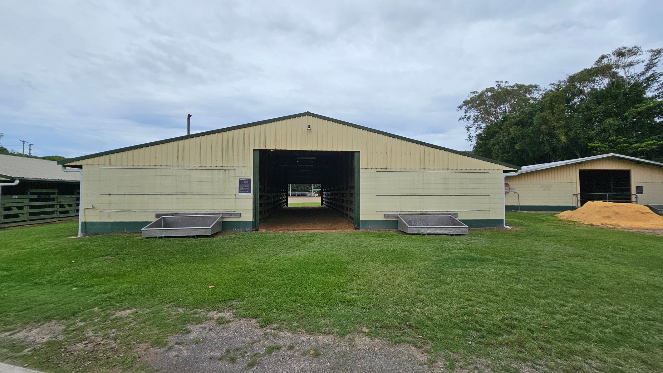 A light yellow barn with a green base stands in the centre, featuring a large open entrance. Two metal troughs are positioned on either side of the entrance. The ground is covered with grass, and a dirt path leads up to the barn. To the right, there's another similar structure with a pile of sand in front. Trees are visible in the background, adding greenery to the scene. A light yellow barn with a green base stands in the centre, featuring a large open entrance. Two metal troughs are positioned on either side of the entrance. The ground is covered with grass, and a dirt path leads up to the barn. To the right, there's another similar structure with a pile of sand in front. Trees are visible in the background, adding greenery to the scene.
