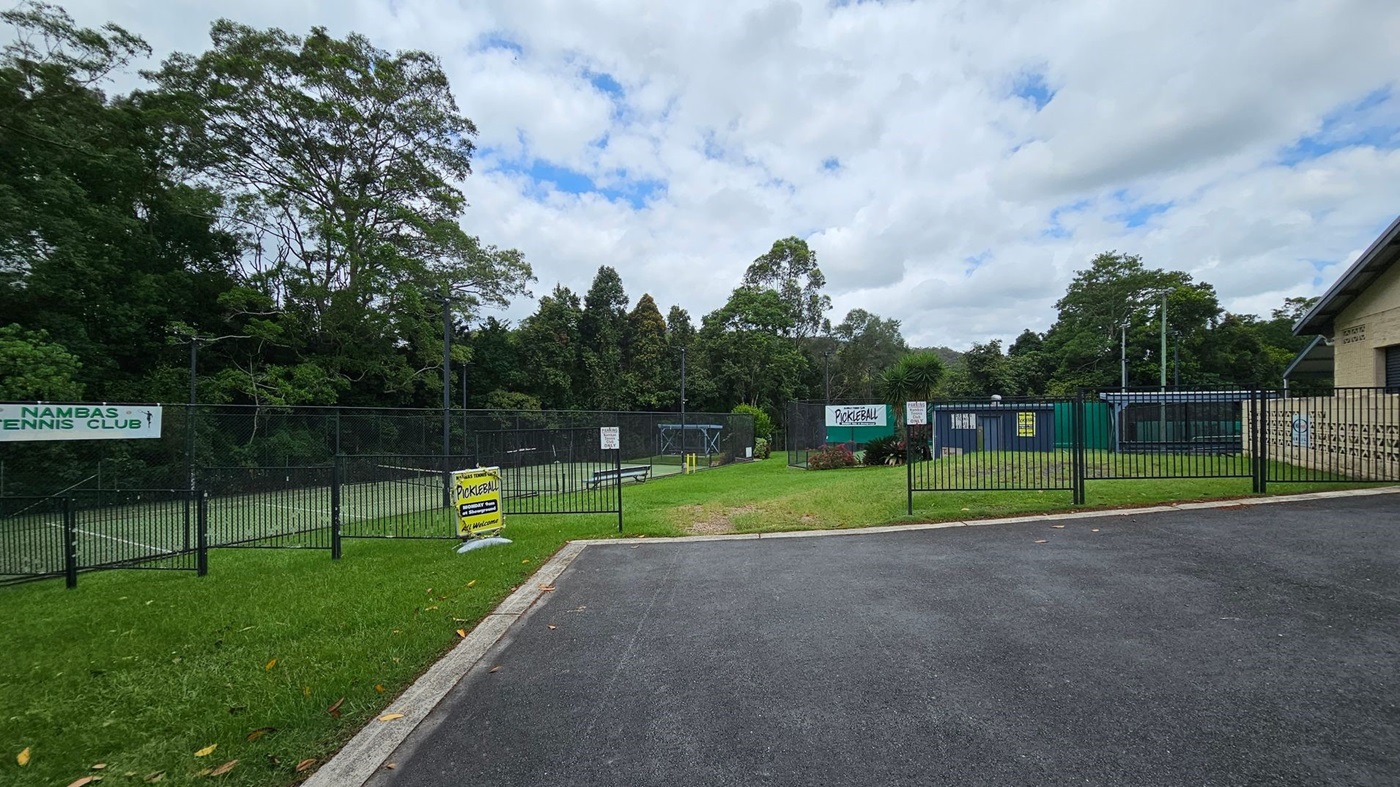 A paved road curves on the right, leading to a building labelled "POULTRY LES DOWDLE PAVILION" with a light blue door. On the left, a tennis court is enclosed by a green fence, with trees in the background. A tree stands in the foreground on a grassy area, with scattered yellow leaves on the ground. A lamppost is next to the tree. A paved road curves on the right, leading to a building labelled "POULTRY LES DOWDLE PAVILION" with a light blue door. On the left, a tennis court is enclosed by a green fence, with trees in the background. A tree stands in the foreground on a grassy area, with scattered yellow leaves on the ground. A lamppost is next to the tree.