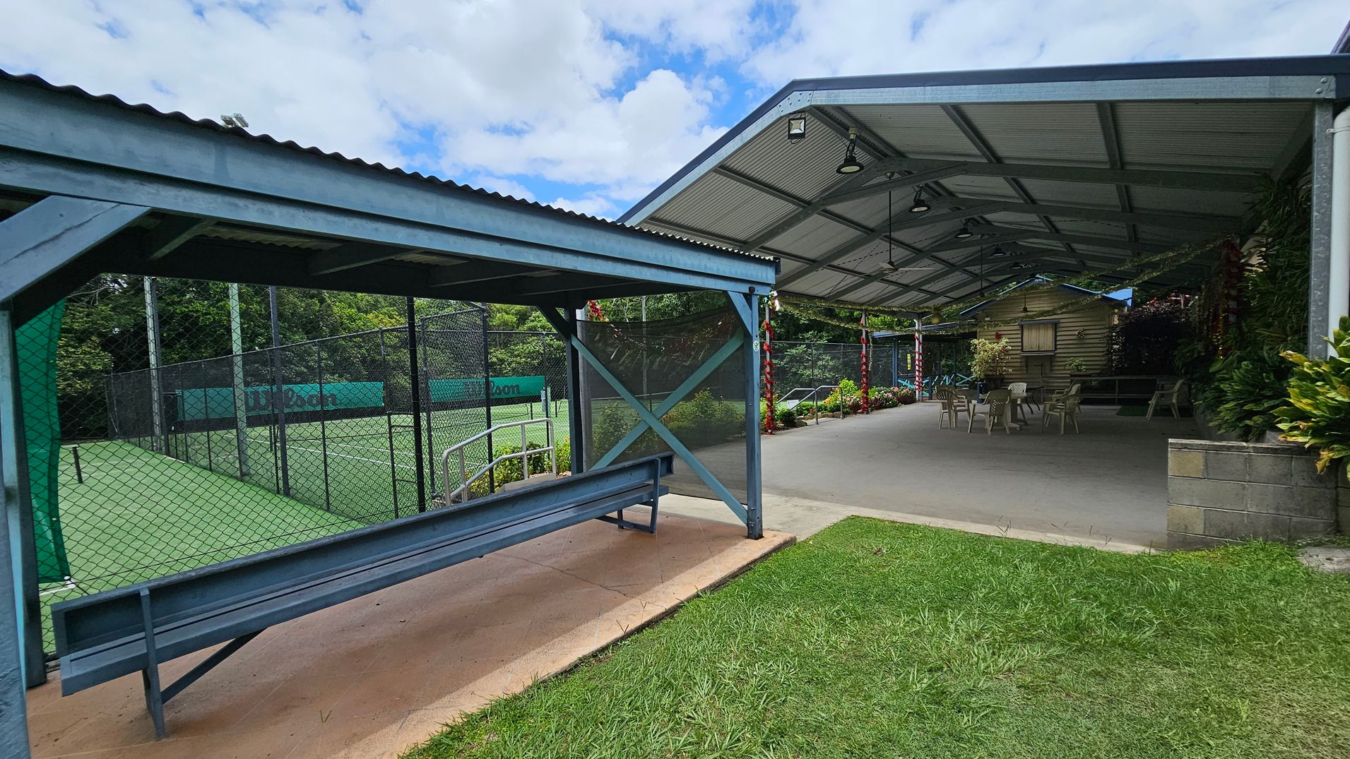 A covered outdoor area with a metal roof and open sides. On the left, there's a fenced tennis court with a green surface and a bench under a small shelter. On the right, a larger covered space with a concrete floor has several plastic chairs and tables. The background features lush greenery and a small wooden structure. The foreground has a patch of grass. A covered outdoor area with a metal roof and open sides. On the left, there's a fenced tennis court with a green surface and a bench under a small shelter. On the right, a larger covered space with a concrete floor has several plastic chairs and tables. The background features lush greenery and a small wooden structure. The foreground has a patch of grass.