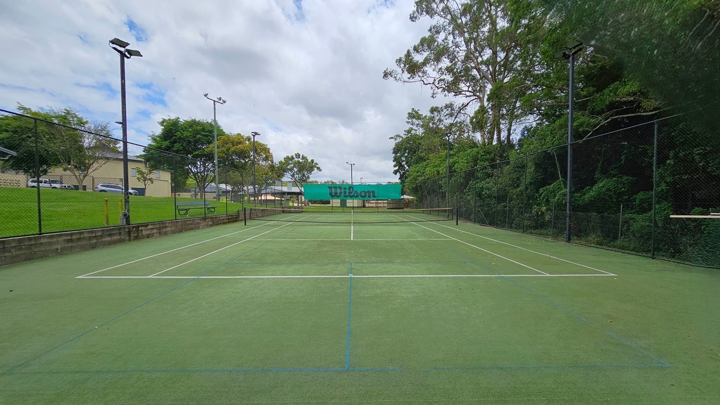 A tennis court with a green surface and white lines. In the centre, a net with a "Wilson" banner. On the left, a grassy area with trees and a building in the background. On the right, dense trees and foliage. Tall floodlights are positioned around the court, and a chain-link fence encloses the area. A tennis court with a green surface and white lines. In the centre, a net with a "Wilson" banner. On the left, a grassy area with trees and a building in the background. On the right, dense trees and foliage. Tall floodlights are positioned around the court, and a chain-link fence encloses the area.