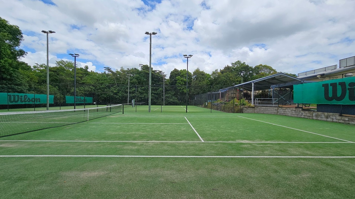 A tennis court with a green artificial grass surface and white lines. The net runs across the middle. On the left and right, green Wilson-branded banners are attached to the fences. Tall floodlights are positioned around the court. In the background, there are trees and a covered seating area with a metal roof and brick base on the right side. A tennis court with a green artificial grass surface and white lines. The net runs across the middle. On the left and right, green Wilson-branded banners are attached to the fences. Tall floodlights are positioned around the court. In the background, there are trees and a covered seating area with a metal roof and brick base on the right side.