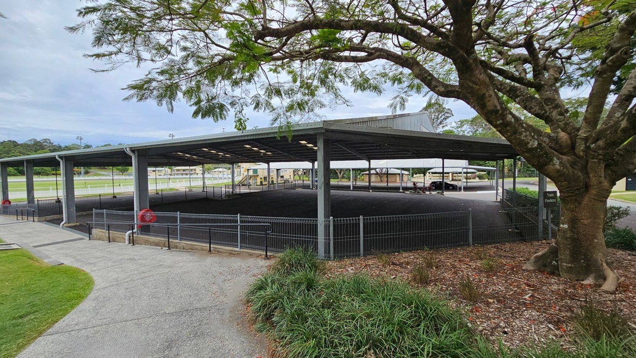 A large, open-sided pavilion with a metal roof and a sign reading "Ray Grace Pavilion" at the front. The structure is supported by metal beams and surrounded by a low hedge. In the background, there are grassy areas and additional buildings. A tree with green foliage is visible on the left. The ground is paved with asphalt, and there are signs and a small path leading to the pavilion. A large, open-sided pavilion with a metal roof and a sign reading "Ray Grace Pavilion" at the front. The structure is supported by metal beams and surrounded by a low hedge. In the background, there are grassy areas and additional buildings. A tree with green foliage is visible on the left. The ground is paved with asphalt, and there are signs and a small path leading to the pavilion.