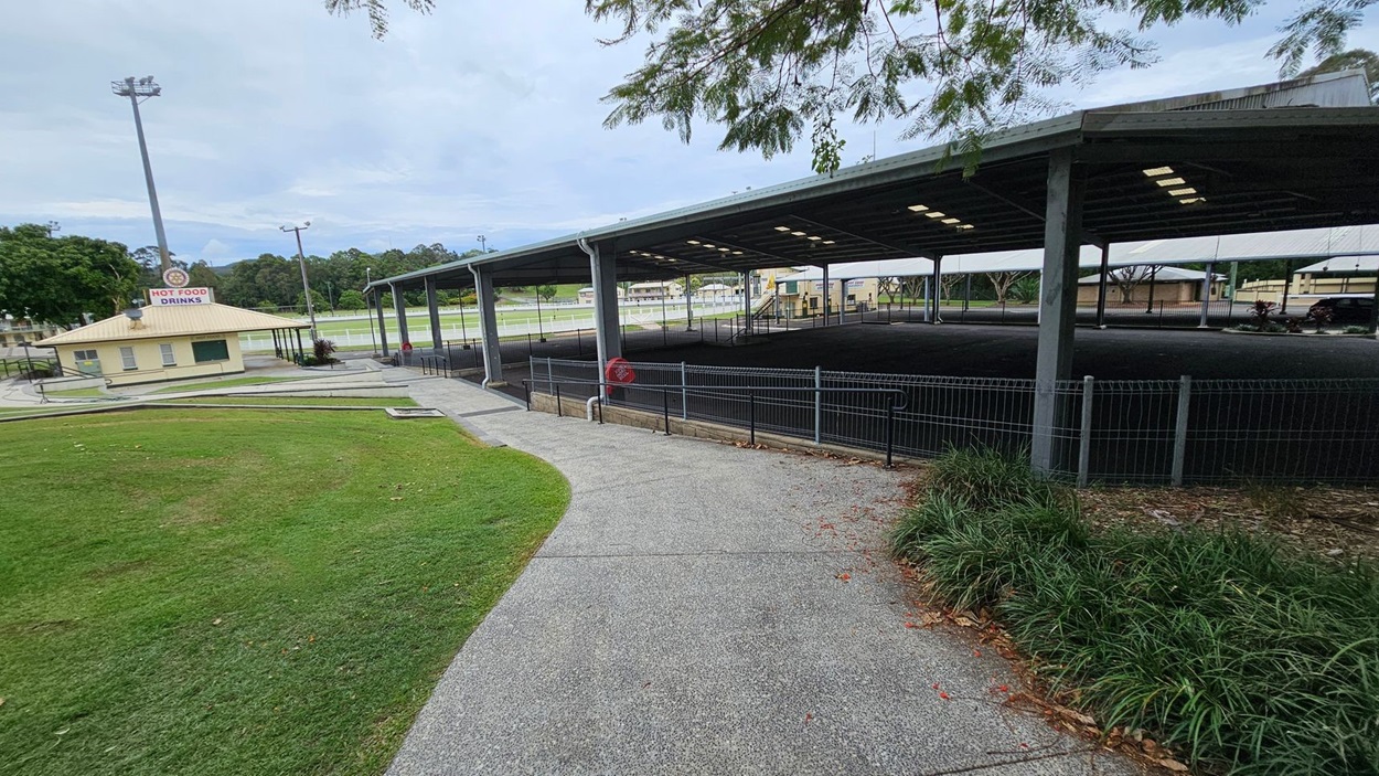 A paved path curves through a grassy area with a small building on the left displaying a "Hot Food Drinks" sign. To the right, a large covered structure with a metal roof and open sides is visible, surrounded by a fence. In the background, there are trees and a sports field. A tall light pole stands near the building, and some foliage is visible at the top edge of the image. A paved path curves through a grassy area with a small building on the left displaying a "Hot Food Drinks" sign. To the right, a large covered structure with a metal roof and open sides is visible, surrounded by a fence. In the background, there are trees and a sports field. A tall light pole stands near the building, and some foliage is visible at the top edge of the image.