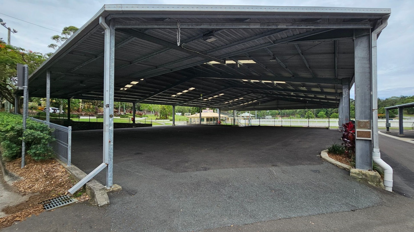 A large, open-sided metal shelter with a corrugated roof supported by metal beams. The ground is paved with asphalt. On the left, there is a fence with greenery behind it. In the background, a small building is visible, surrounded by trees and a grassy area. On the right, a small garden bed with red and green plants is next to a drainage pipe. The ceiling has several skylights allowing natural light to enter. A large, open-sided metal shelter with a corrugated roof supported by metal beams. The ground is paved with asphalt. On the left, there is a fence with greenery behind it. In the background, a small building is visible, surrounded by trees and a grassy area. On the right, a small garden bed with red and green plants is next to a drainage pipe. The ceiling has several skylights allowing natural light to enter.