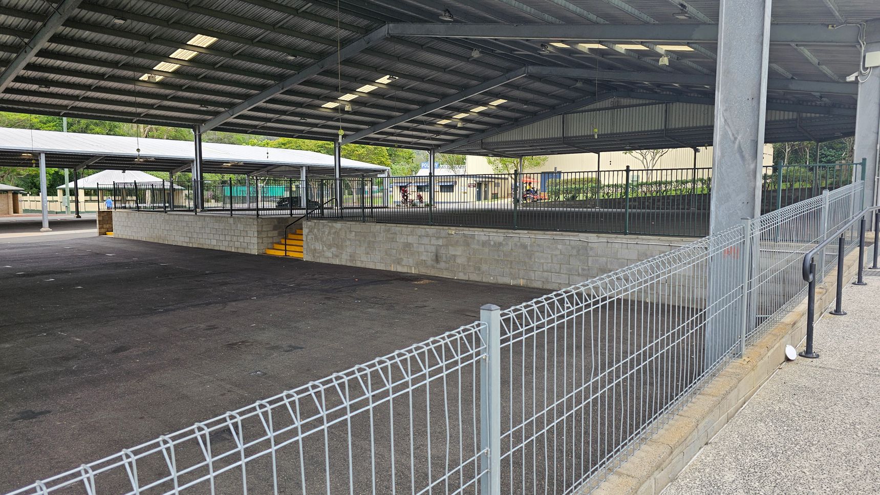 A large, open-sided structure with a metal roof and several support beams. The floor is paved with dark asphalt. In the centre, there is a raised platform with a low brick wall and yellow steps leading up to it. Metal fencing surrounds the platform. In the background, there are trees and a building with a sign partially visible. A wire fence runs along the right side, with a handrail attached to a low wall. A large, open-sided structure with a metal roof and several support beams. The floor is paved with dark asphalt. In the centre, there is a raised platform with a low brick wall and yellow steps leading up to it. Metal fencing surrounds the platform. In the background, there are trees and a building with a sign partially visible. A wire fence runs along the right side, with a handrail attached to a low wall.
