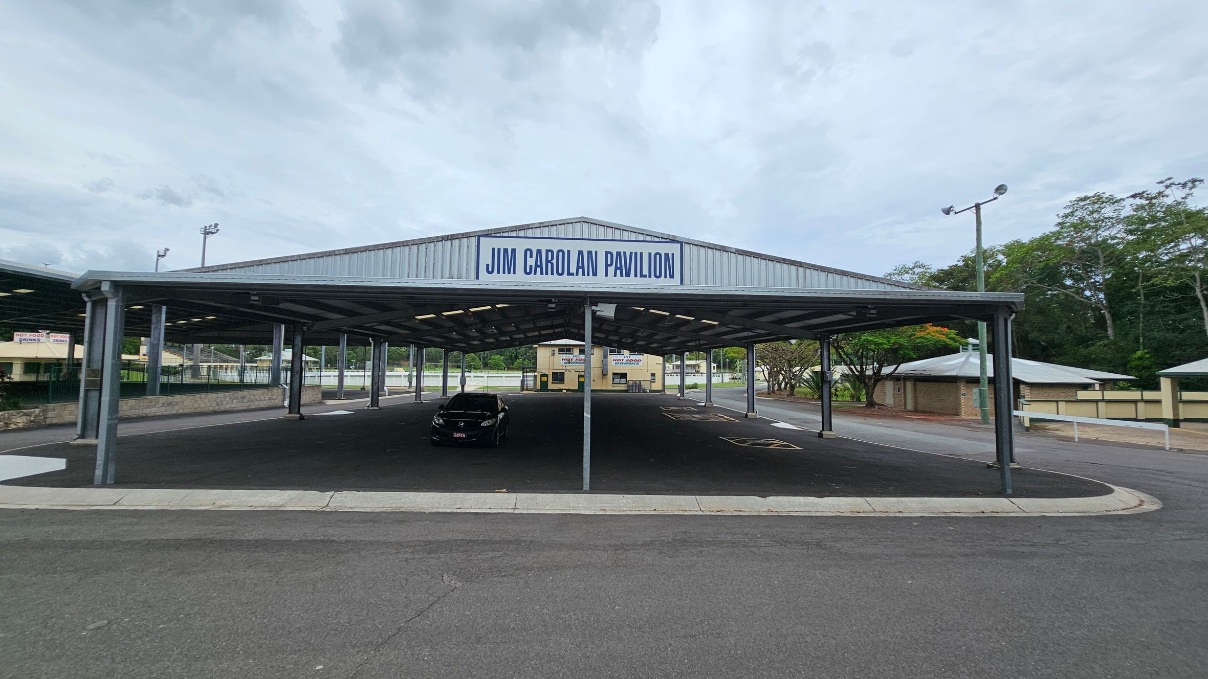 A large, open-sided pavilion with a metal roof labelled "Jim Carolan Pavilion" at the top. Underneath, a single black car is parked on the dark asphalt surface. In the background, there are trees and a few small buildings, including a yellow one with signs. The area is surrounded by a low fence and there are streetlights on tall poles. The ground is paved, and the pavilion is supported by metal columns. A large, open-sided pavilion with a metal roof labelled "Jim Carolan Pavilion" at the top. Underneath, a single black car is parked on the dark asphalt surface. In the background, there are trees and a few small buildings, including a yellow one with signs. The area is surrounded by a low fence and there are streetlights on tall poles. The ground is paved, and the pavilion is supported by metal columns.