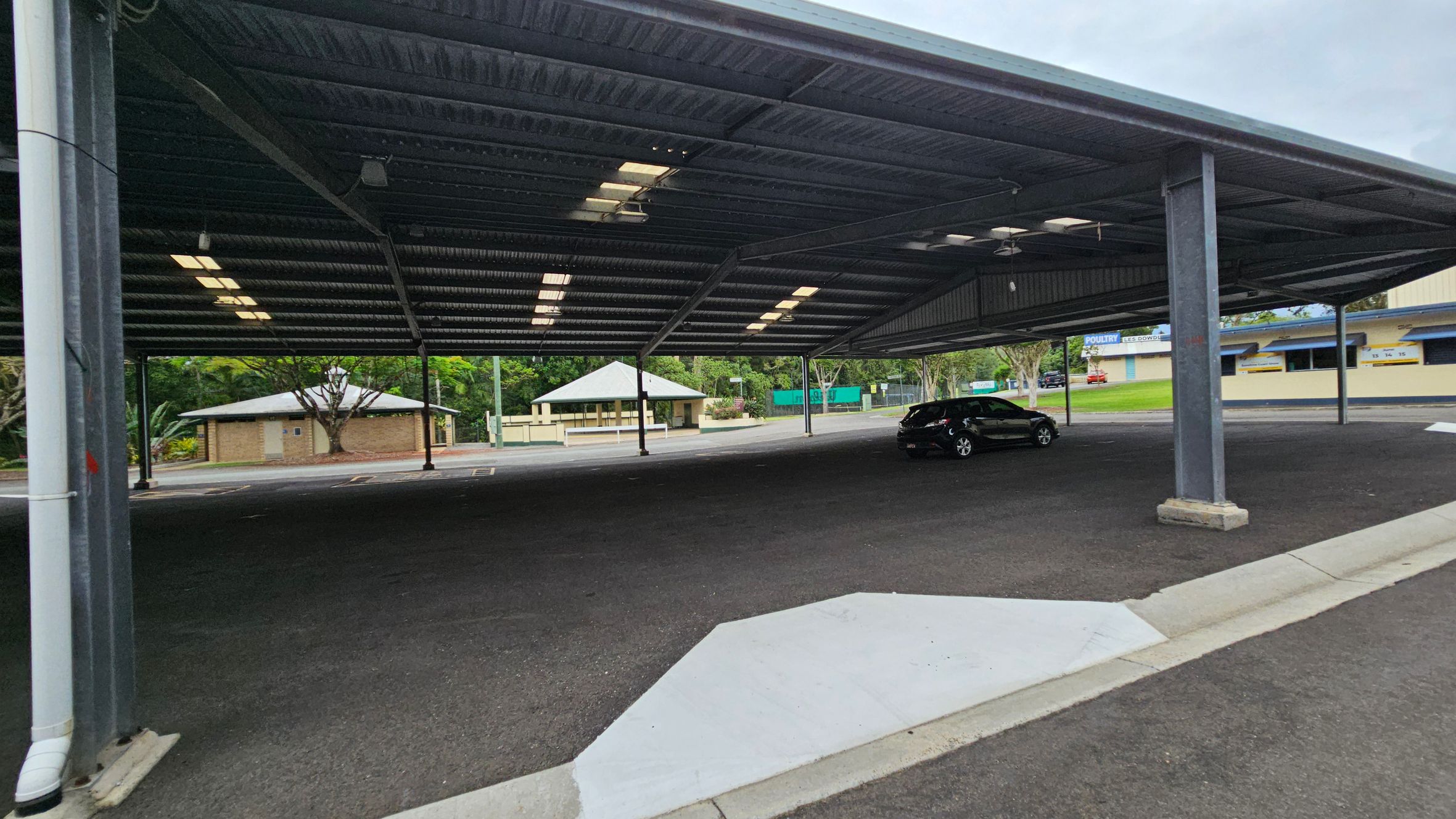 A covered parking area with a dark asphalt floor and a metal roof supported by columns. A single black car is parked on the right side. In the background, there are small buildings with light-coloured walls and green roofs, surrounded by trees and greenery. The ceiling has rows of fluorescent lights. A covered parking area with a dark asphalt floor and a metal roof supported by columns. A single black car is parked on the right side. In the background, there are small buildings with light-coloured walls and green roofs, surrounded by trees and greenery. The ceiling has rows of fluorescent lights.