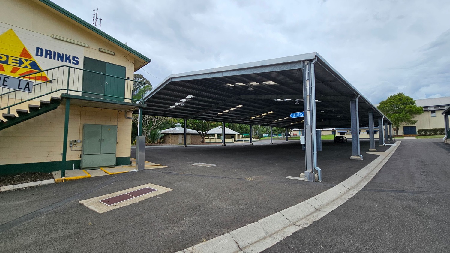 A paved area with a covered structure supported by metal beams on the right. A sign indicating "TOILETS" hangs from the structure. On the left, a building with a staircase and a sign that reads "DRINKS" is visible. In the background, there are trees and small buildings with green roofs. The ground is asphalt, and the area appears to be part of a larger complex or park. A paved area with a covered structure supported by metal beams on the right. A sign indicating "TOILETS" hangs from the structure. On the left, a building with a staircase and a sign that reads "DRINKS" is visible. In the background, there are trees and small buildings with green roofs. The ground is asphalt, and the area appears to be part of a larger complex or park.