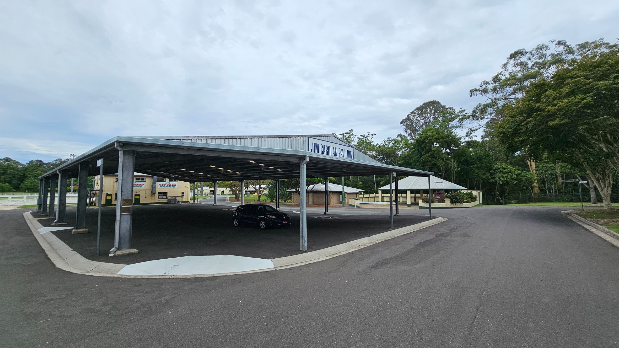 A large open pavilion with a metal roof labelled "Jim Carolan Pavilion" is on the left. Underneath, a black car is parked. To the right, a paved road curves around the structure, bordered by trees and greenery. In the background, there are small buildings, including a food stand with signs for "Hot Food" and "Cold Drinks". The area is surrounded by trees and vegetation. A large open pavilion with a metal roof labelled "Jim Carolan Pavilion" is on the left. Underneath, a black car is parked. To the right, a paved road curves around the structure, bordered by trees and greenery. In the background, there are small buildings, including a food stand with signs for "Hot Food" and "Cold Drinks". The area is surrounded by trees and vegetation.