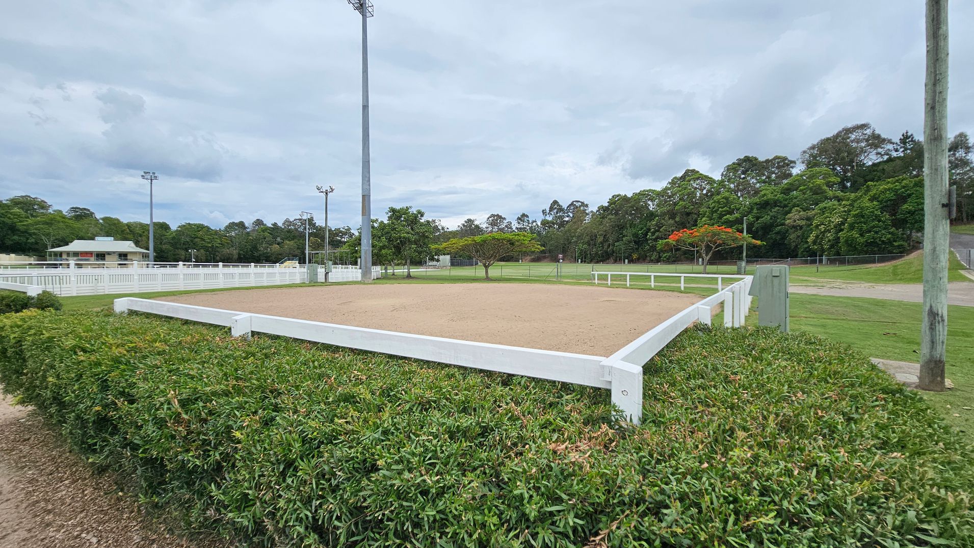 A rectangular sandy area is surrounded by a white fence, with green hedges in the foreground. In the background, there are tall trees and a few scattered smaller trees with red flowers. A building with a green roof is visible to the left, and several tall floodlights are positioned around the area. The ground is grassy, and a wooden utility pole stands on the right. A rectangular sandy area is surrounded by a white fence, with green hedges in the foreground. In the background, there are tall trees and a few scattered smaller trees with red flowers. A building with a green roof is visible to the left, and several tall floodlights are positioned around the area. The ground is grassy, and a wooden utility pole stands on the right.