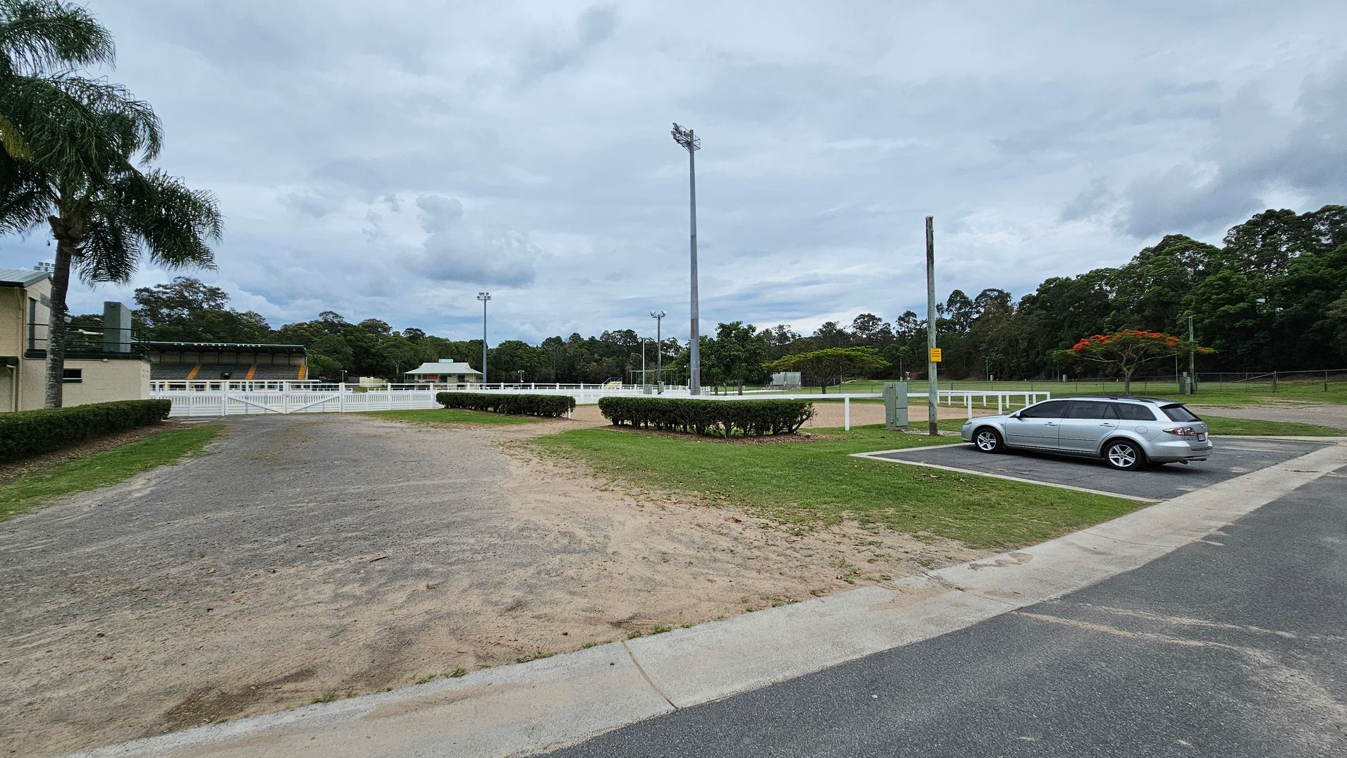 A sports field with a large light pole in the centre. On the left, there's a building with a palm tree nearby. A white fence surrounds the field, and there are hedges along the path. On the right, a silver car is parked in a designated parking area. The background features trees and additional light poles. The ground is a mix of grass and dirt, with a paved road in the foreground. A sports field with a large light pole in the centre. On the left, there's a building with a palm tree nearby. A white fence surrounds the field, and there are hedges along the path. On the right, a silver car is parked in a designated parking area. The background features trees and additional light poles. The ground is a mix of grass and dirt, with a paved road in the foreground.