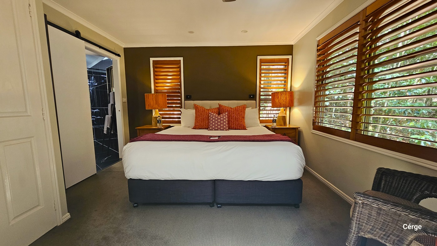 A queen-size bed occupies the center, adorned with tasteful decor. On either side, table lamp shades in coordinating colours with the bed linens illuminate the space. Three windows provide natural light, one large window to the right and two smaller ones behind the bed headrest, each adorned with brown window blinds. To the left, a glimpse of the accessible bathroom is visible, while to the right, a rattan-designed chair is positioned. The ceiling and walls are painted in a pristine white hue. A queen-size bed occupies the center, adorned with tasteful decor. On either side, table lamp shades in coordinating colours with the bed linens illuminate the space. Three windows provide natural light, one large window to the right and two smaller ones behind the bed headrest, each adorned with brown window blinds. To the left, a glimpse of the accessible bathroom is visible, while to the right, a rattan-designed chair is positioned. The ceiling and walls are painted in a pristine white hue.