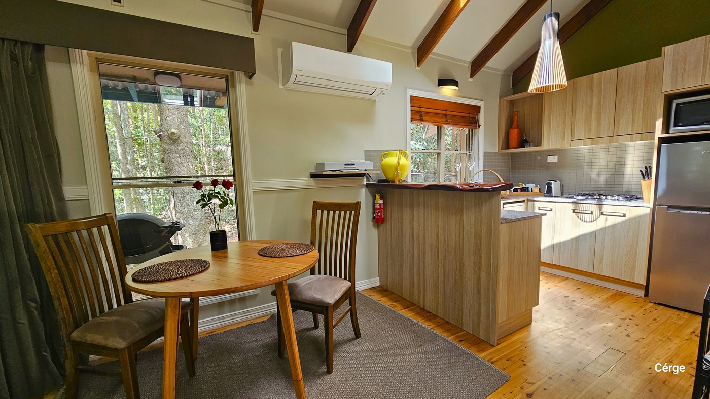 In the Dove Cottage kitchen, timber flooring supports a round table with two chairs, adorned with a flower vase and table mats. A glass window offers a view of the rainforest, complemented by an olive curtain. To the right, a tiled kitchen sink, fridge, stove, and accessories are arranged. There is also a mini kitchen island near the table. Wall-mounted air conditioning and lighting fixtures illuminate the space, while the ceiling features a wooden plank design with a hanging light fixture. In the Dove Cottage kitchen, timber flooring supports a round table with two chairs, adorned with a flower vase and table mats. A glass window offers a view of the rainforest, complemented by an olive curtain. To the right, a tiled kitchen sink, fridge, stove, and accessories are arranged. There is also a mini kitchen island near the table. Wall-mounted air conditioning and lighting fixtures illuminate the space, while the ceiling features a wooden plank design with a hanging light fixture.
