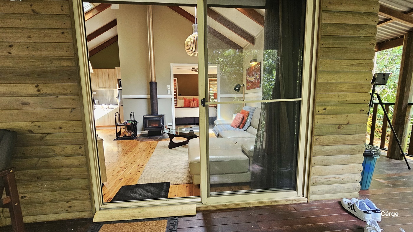 In this view, a glass sliding door serves as the entrance to the cottage. Both the interior and exterior floors are made of timber. Inside, a cream leather couch adorned with pillows and a glass center table are present. On the right wall, there is a framed decoration and a hanging light fixture from the ceiling. A mini shelf displays magazines and chopped wood for decoration. Outside, on the verandah, a cushioned chair is placed, with floor mats positioned before the door entrance. In this view, a glass sliding door serves as the entrance to the cottage. Both the interior and exterior floors are made of timber. Inside, a cream leather couch adorned with pillows and a glass center table are present. On the right wall, there is a framed decoration and a hanging light fixture from the ceiling. A mini shelf displays magazines and chopped wood for decoration. Outside, on the verandah, a cushioned chair is placed, with floor mats positioned before the door entrance.