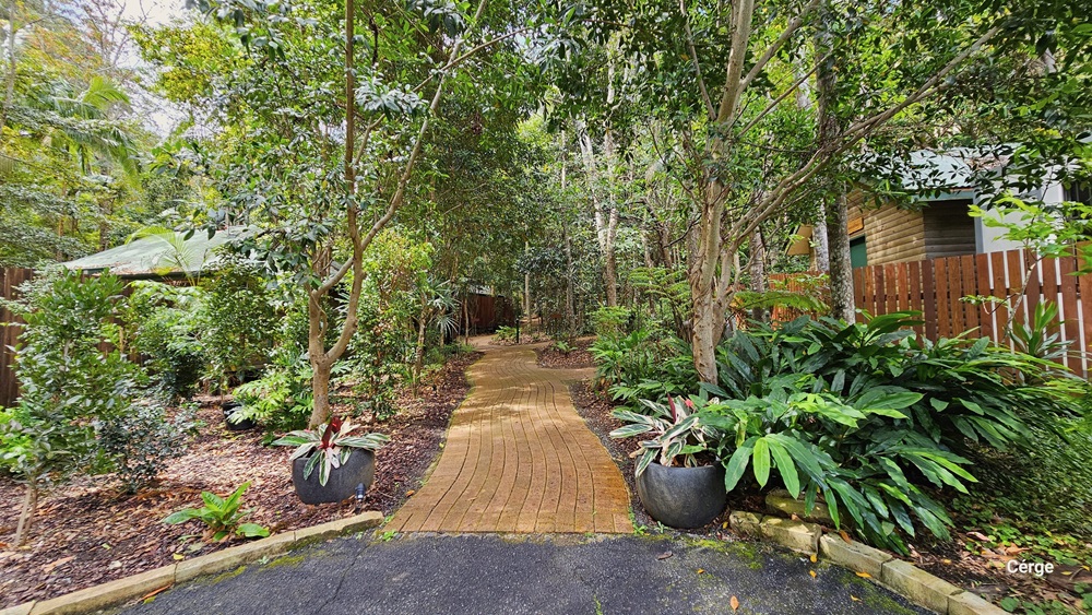 Approaching Kookaburra Cottage, to the right of the road is a shaded carport where a red car is parked. A mini trolley stands nearby. On the left, a plant box bears a signage reminder. Further to the left, there is a row of shaded carports. Tall trees, shrubs, plants, and grass surround the area. Approaching Kookaburra Cottage, to the right of the road is a shaded carport where a red car is parked. A mini trolley stands nearby. On the left, a plant box bears a signage reminder. Further to the left, there is a row of shaded carports. Tall trees, shrubs, plants, and grass surround the area.