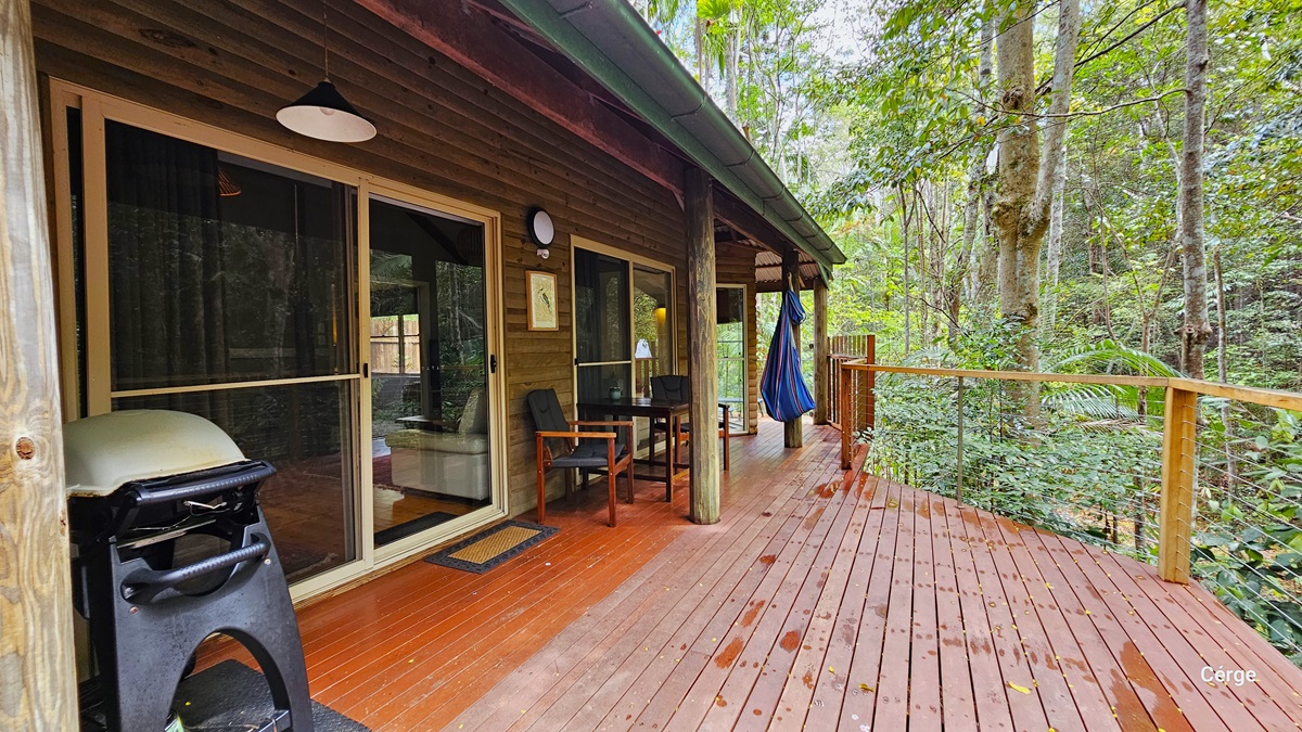This is the verandah serving as the entrance to the cottage. The building features a wooden plank design, and the floor is constructed with timber. A metal railing secures the area. In this view, large trees are visible on the right. On the left, a sliding door is present with a floor mat placed before it. A cushioned chair and table are positioned to the right of the door. To the left of the door, a barbecue grill is located. Lighting fixtures and wooden frame decorations are also visible. This is the verandah serving as the entrance to the cottage. The building features a wooden plank design, and the floor is constructed with timber. A metal railing secures the area. In this view, large trees are visible on the right. On the left, a sliding door is present with a floor mat placed before it. A cushioned chair and table are positioned to the right of the door. To the left of the door, a barbecue grill is located. Lighting fixtures and wooden frame decorations are also visible.