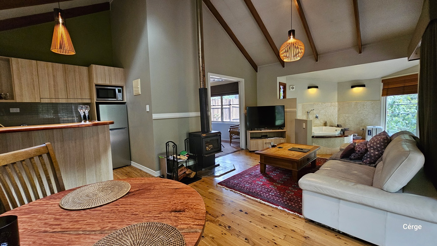 To the right is a grey leather couch adorned with pillows, accompanied by a wooden center table. To the right, a flatscreen TV is placed on top of a TV rack. Next to it is a wash basin. A glass door on the right leads to the pool. The floor is made of timber, and a large red carpet embellishes the living room. To the left is a round table with two place mats, near the fully furnished kitchen area. Uniquely designed light fixtures hang from the wooden plank design ceiling. To the right is a grey leather couch adorned with pillows, accompanied by a wooden center table. To the right, a flatscreen TV is placed on top of a TV rack. Next to it is a wash basin. A glass door on the right leads to the pool. The floor is made of timber, and a large red carpet embellishes the living room. To the left is a round table with two place mats, near the fully furnished kitchen area. Uniquely designed light fixtures hang from the wooden plank design ceiling.