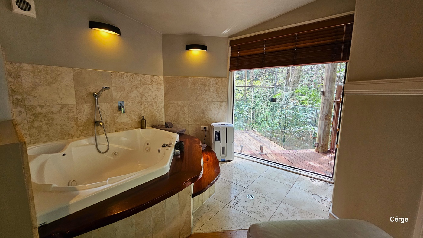 Inside the bathroom, there is a white bathtub fixed against the wall, accompanied by accessible two-step wooden stairs. The bathroom area is painted cream, and the wall features marble design tiles. Lighting fixtures are affixed to the left wall, and a portable heater is placed in the corner. From this vantage point, a view of the forest green outside is also visible. Inside the bathroom, there is a white bathtub fixed against the wall, accompanied by accessible two-step wooden stairs. The bathroom area is painted cream, and the wall features marble design tiles. Lighting fixtures are affixed to the left wall, and a portable heater is placed in the corner. From this vantage point, a view of the forest green outside is also visible.