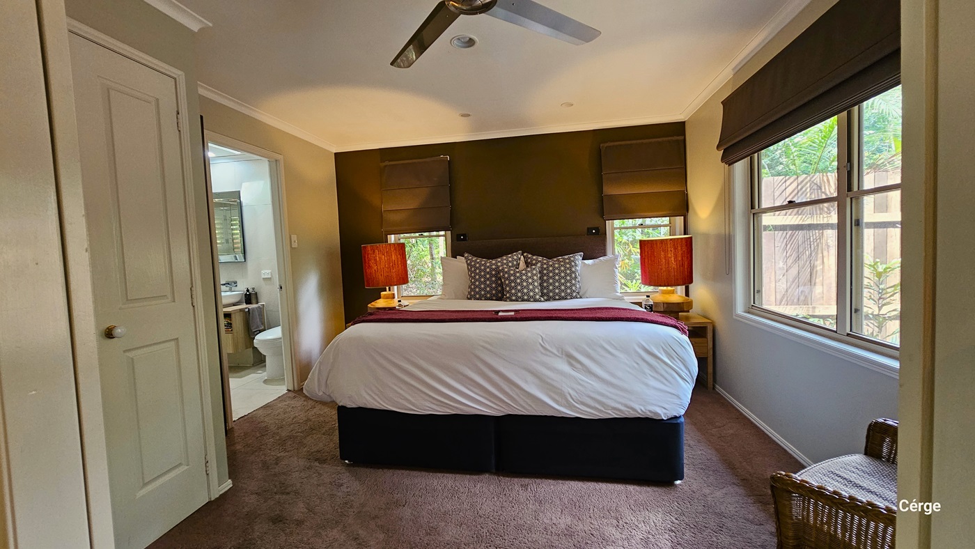 In the middle of the room, a large bed is neatly adorned with pillows and bed linens. Wooden-colored lamps on two side tables flank the bed. To the right, there's a sliding window, and two smaller windows above the bed, all equipped with thick brown blinds. In the left corner, a cushioned rattan chair is placed, and a glimpse of the bathroom is visible. The floor is covered with a brown furry carpet, and a ceiling fan is installed on the white ceiling. In the middle of the room, a large bed is neatly adorned with pillows and bed linens. Wooden-colored lamps on two side tables flank the bed. To the right, there's a sliding window, and two smaller windows above the bed, all equipped with thick brown blinds. In the left corner, a cushioned rattan chair is placed, and a glimpse of the bathroom is visible. The floor is covered with a brown furry carpet, and a ceiling fan is installed on the white ceiling.