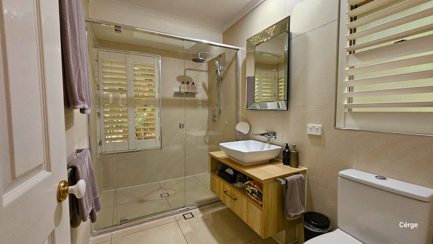 In the bathroom, the toilet is situated near the door on the right, accompanied by a black rubbish bin and a wash basin. A bathroom organizer is positioned underneath the wash basin, holding bathroom essentials. On the wall, a mirror is affixed, along with a small round mirror. Next to it, a glass door encloses the shower area, and windows with blinds are installed on both sides. On the left, a rack holding towels is in place. The bathroom is tiled in cream, and the walls and ceiling are white. In the bathroom, the toilet is situated near the door on the right, accompanied by a black rubbish bin and a wash basin. A bathroom organizer is positioned underneath the wash basin, holding bathroom essentials. On the wall, a mirror is affixed, along with a small round mirror. Next to it, a glass door encloses the shower area, and windows with blinds are installed on both sides. On the left, a rack holding towels is in place. The bathroom is tiled in cream, and the walls and ceiling are white.