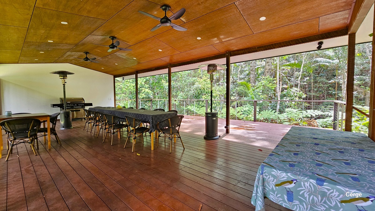 The Rainforest Deck is a spacious area with brown timber plank flooring and a slightly pitched wooden brown roof. Three ceiling fans and a portable heater are installed. Tables and chairs are arranged throughout, including one with a blue tablecloth featuring birds and foliage design. Towards the back, an open verandah secured by metal railings provides views of the surrounding rainforest. The Rainforest Deck is a spacious area with brown timber plank flooring and a slightly pitched wooden brown roof. Three ceiling fans and a portable heater are installed. Tables and chairs are arranged throughout, including one with a blue tablecloth featuring birds and foliage design. Towards the back, an open verandah secured by metal railings provides views of the surrounding rainforest.