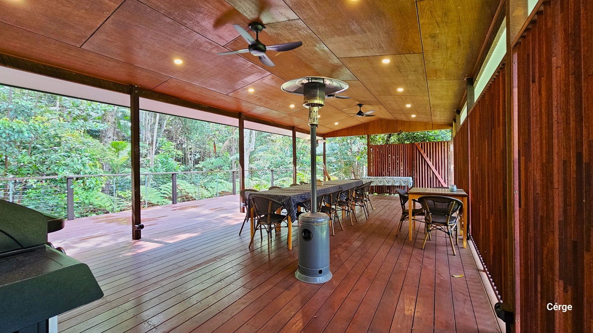 The Rainforest Deck is a spacious area with brown timber plank flooring and a slightly pitched wooden brown roof. Three ceiling fans and a portable heater provide comfort. Tables and chairs are arranged to the sides and the middle. A wall with a wooden plank design extends to the right towards the back. On the left, there's an open verandah secured by metal railings offering views of the surrounding rainforest. The Rainforest Deck is a spacious area with brown timber plank flooring and a slightly pitched wooden brown roof. Three ceiling fans and a portable heater provide comfort. Tables and chairs are arranged to the sides and the middle. A wall with a wooden plank design extends to the right towards the back. On the left, there's an open verandah secured by metal railings offering views of the surrounding rainforest.